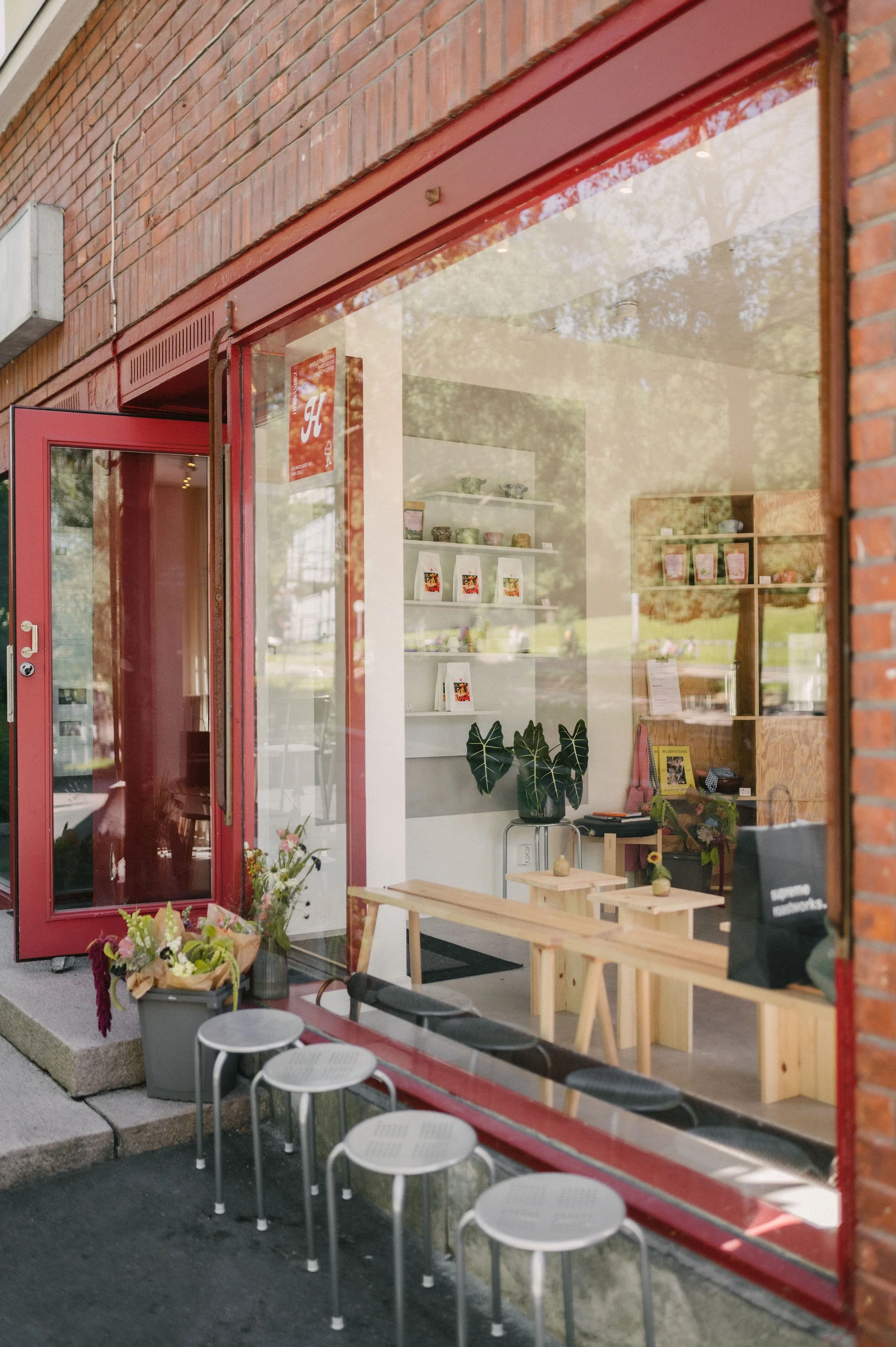 Utsiden av en butikk med store vinduer og rød dørramme. Det er blomster og planter utenfor, og utendørs sitteplasser med tre benker og sirkulære hvite stoler.