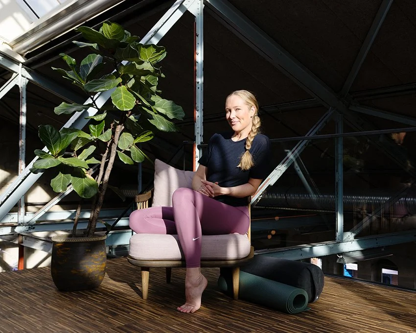 En kvinne i sportslig antrekk som sitter på en lenestol ved et plantestativ i et moderne, industrielt innendørsmiljø.
