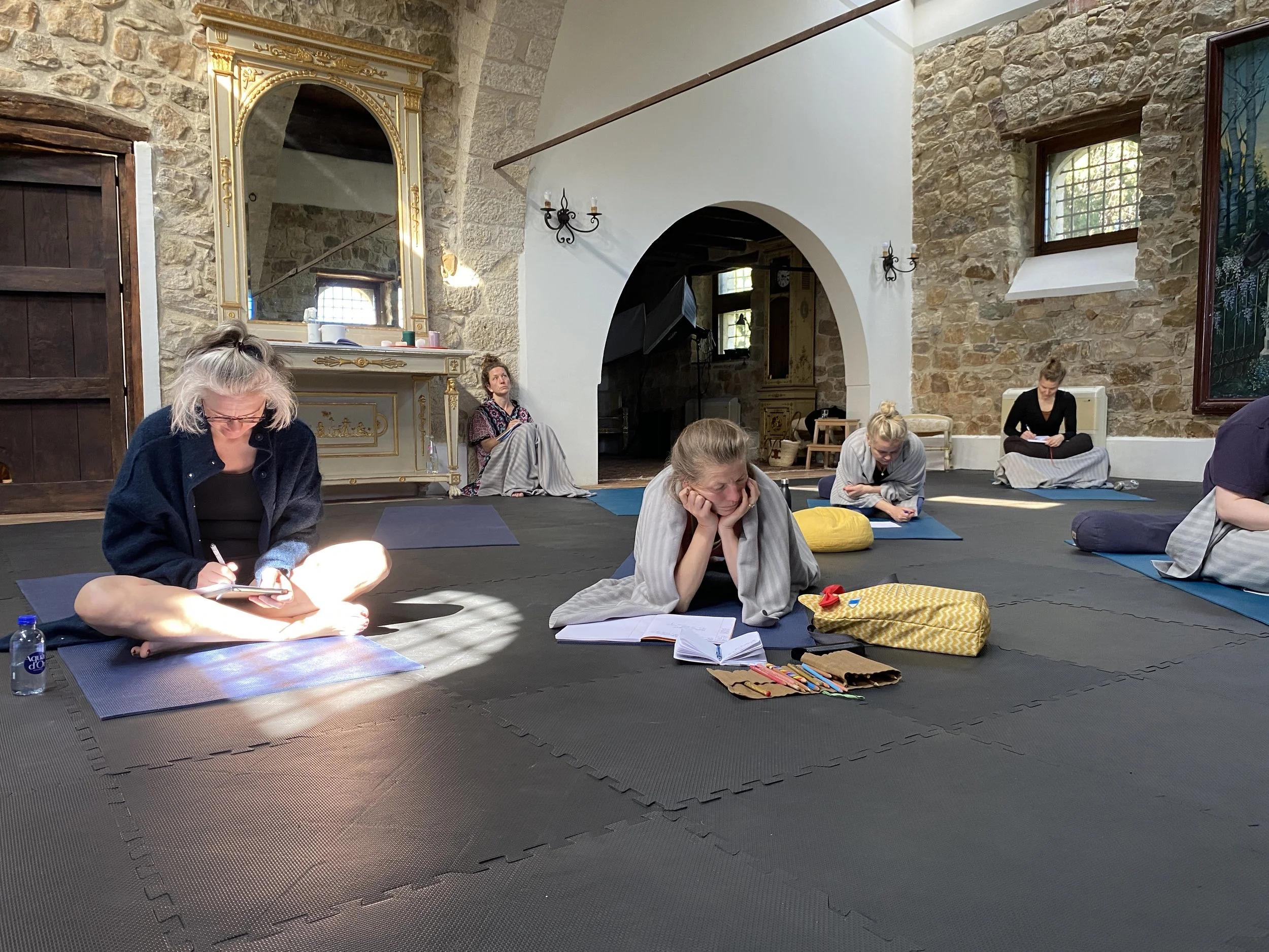 People sitting on mats in a rustic room, engaged in a contemplative or meditative activity, with one person writing, another reading from a book, and others sitting quietly, some with their eyes closed or resting their heads on their hands.