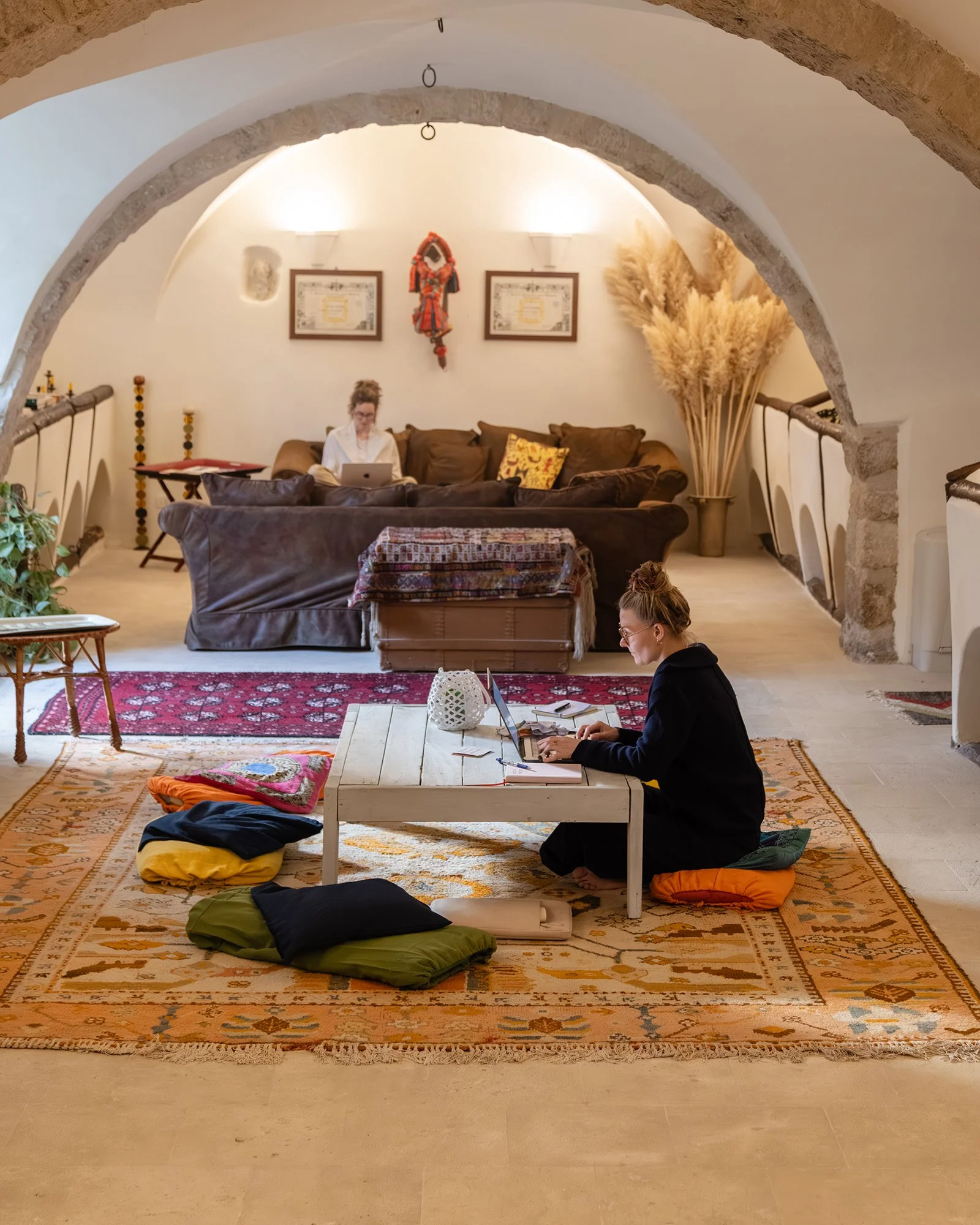 Two women working on laptops in a cozy living room with arched ceiling, decorated with books, plants, and framed art on the wall.