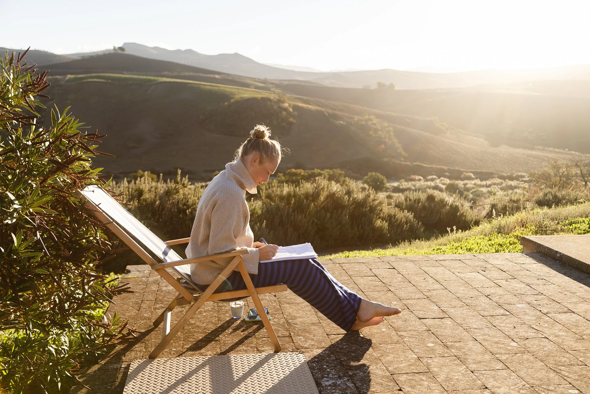 Woman with blonde hair tied up, sitting on a wooden lounge chair on a brick patio, wearing a beige sweater and striped pants, writing in a notebook, with a glass of water and a book beside her, overlooking a scenic mountain and valley landscape at sunset.