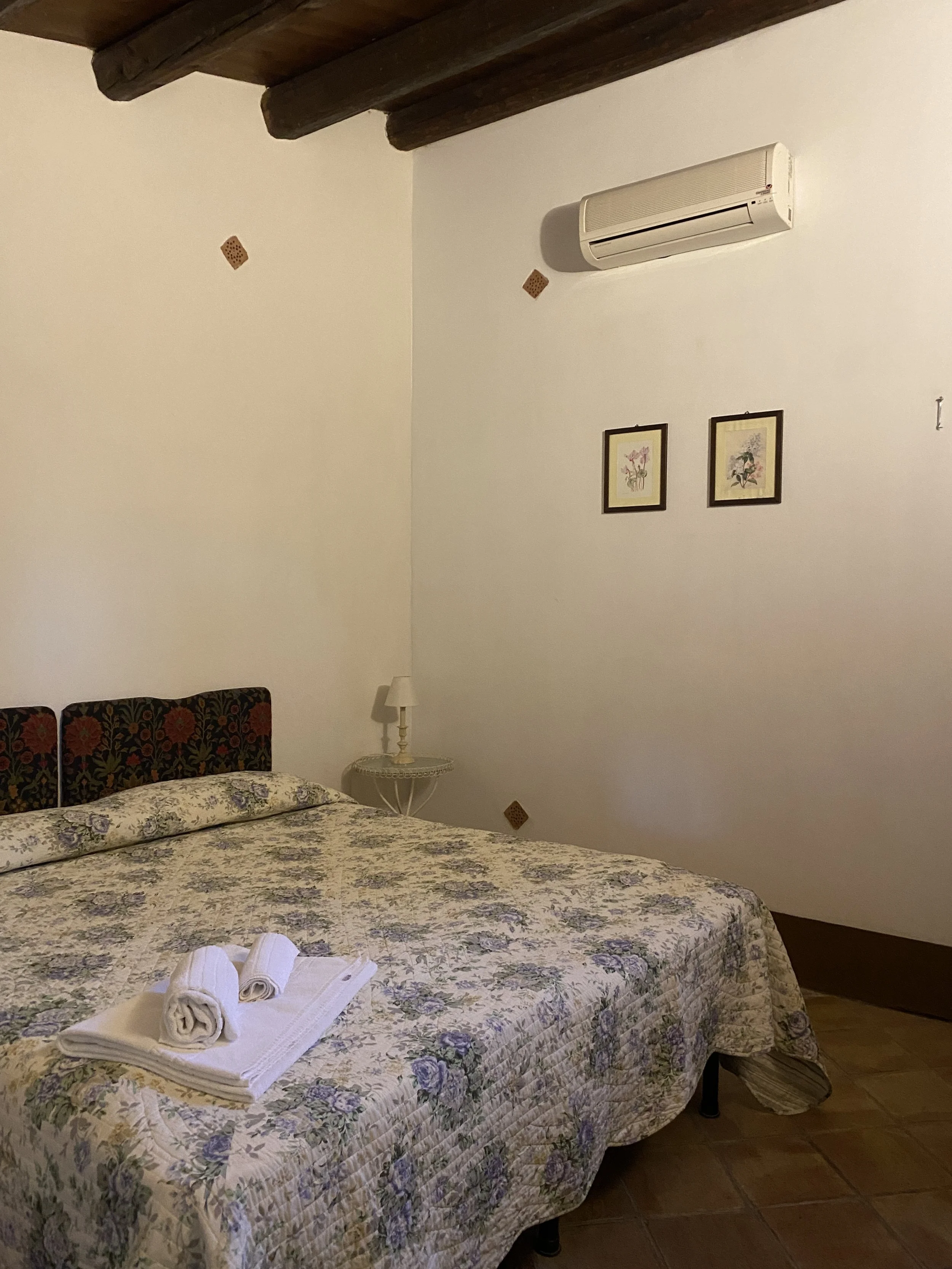 A bedroom with a floral bedspread, two rolled towels on the bed, a small side table with a white lamp, framed botanical prints on the wall, and a wall-mounted air conditioner. The ceiling has wooden beams.