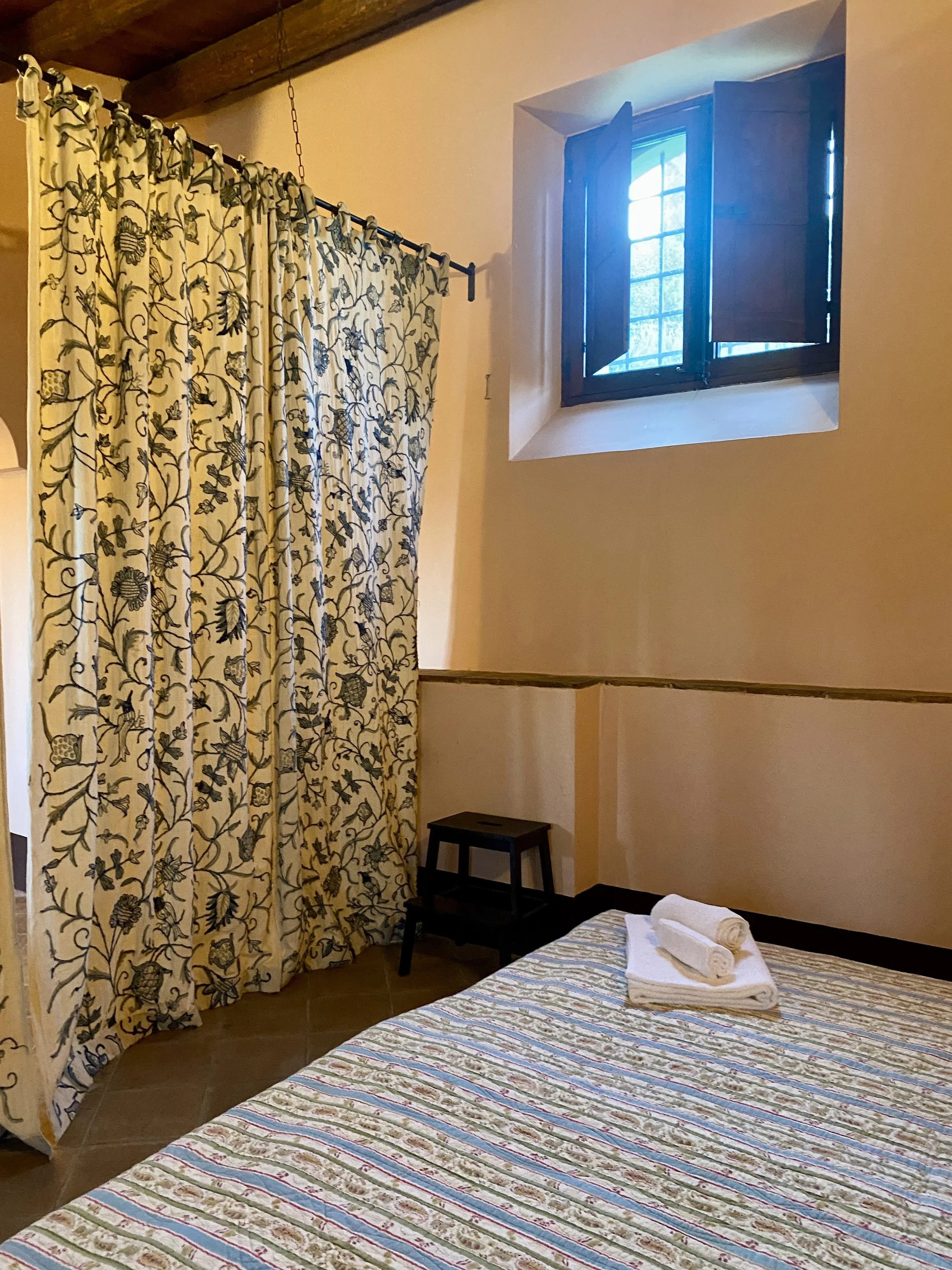 A cozy bedroom with a bed covered in a patterned bedspread, neatly folded towels on top, a small black bedside table, a window with wooden shutters, and a floral curtain hanging from a rod.