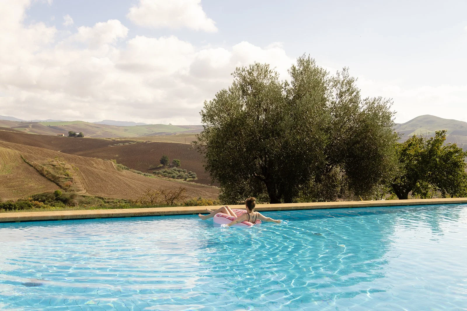 A woman relaxing on a pink float in a swimming pool with a scenic countryside view of rolling hills and trees in the background.
