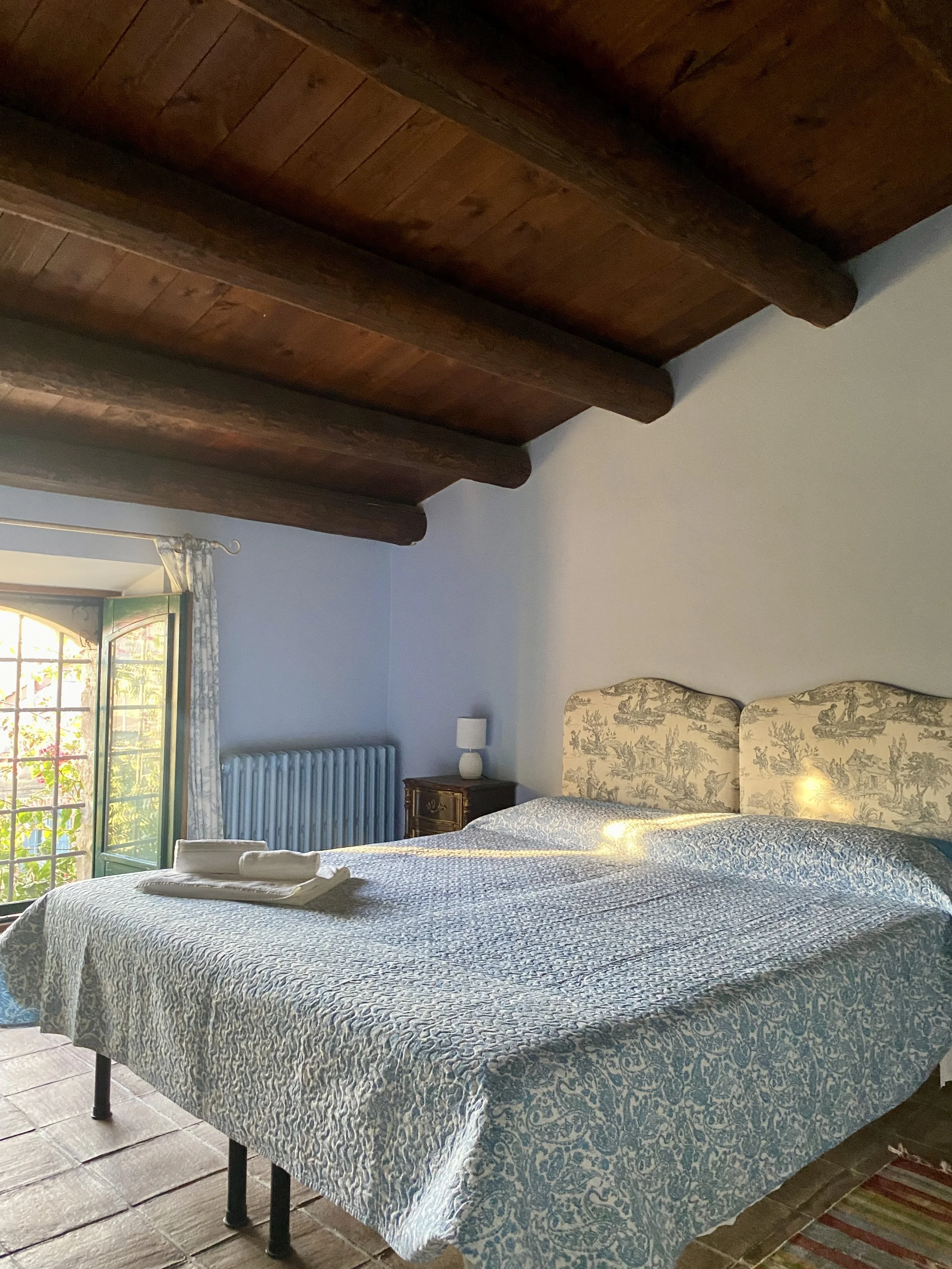 A cozy bedroom featuring a large bed with a blue and white patterned bedspread, a headboard with a pastoral scene design, a small bedside table with a lamp, a radiator, and a window with glass doors and floral curtains, showing a sunny outdoor view.