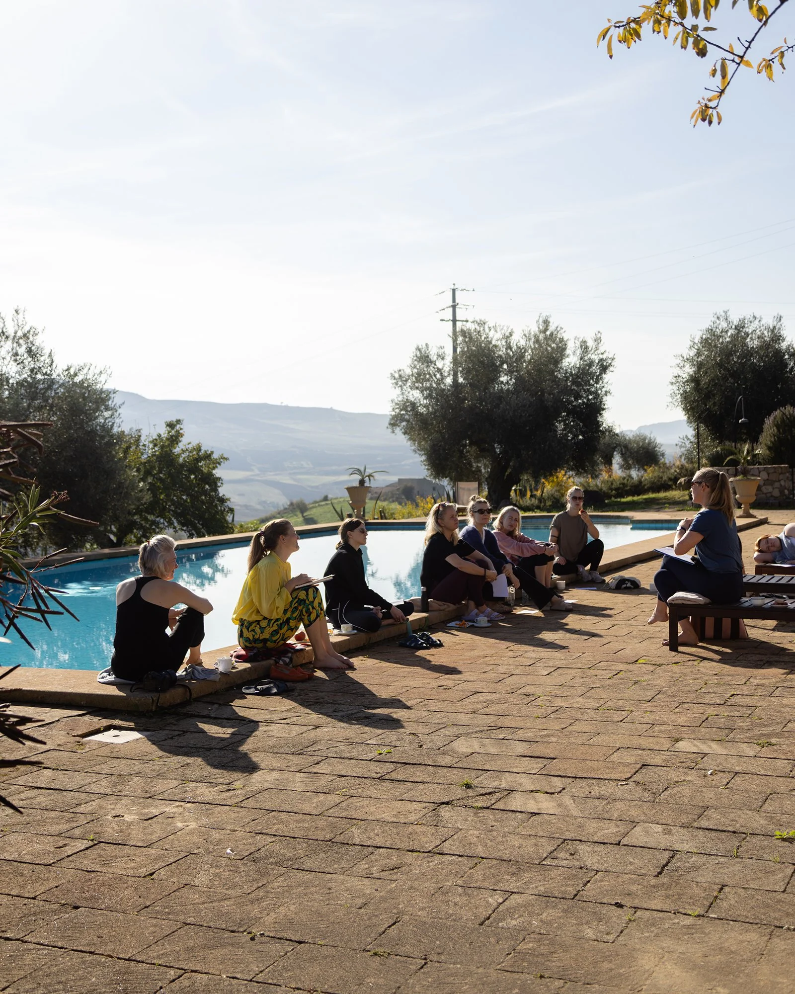 A group of people sitting outside by a pool, engaging in a discussion or class, with a woman leading and a scenic landscape with trees and mountains in the background.
