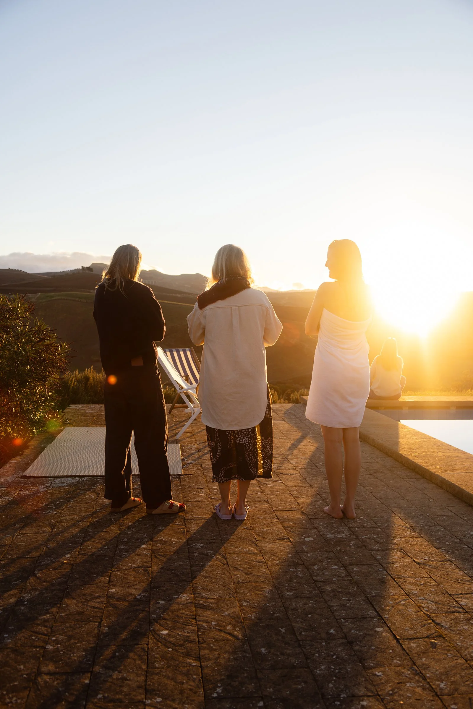 Three women standing on a patio near a pool, watching a sunset in a mountainous landscape. One woman is wrapped in a towel, another in a skirt and jacket, and the third in a towel.