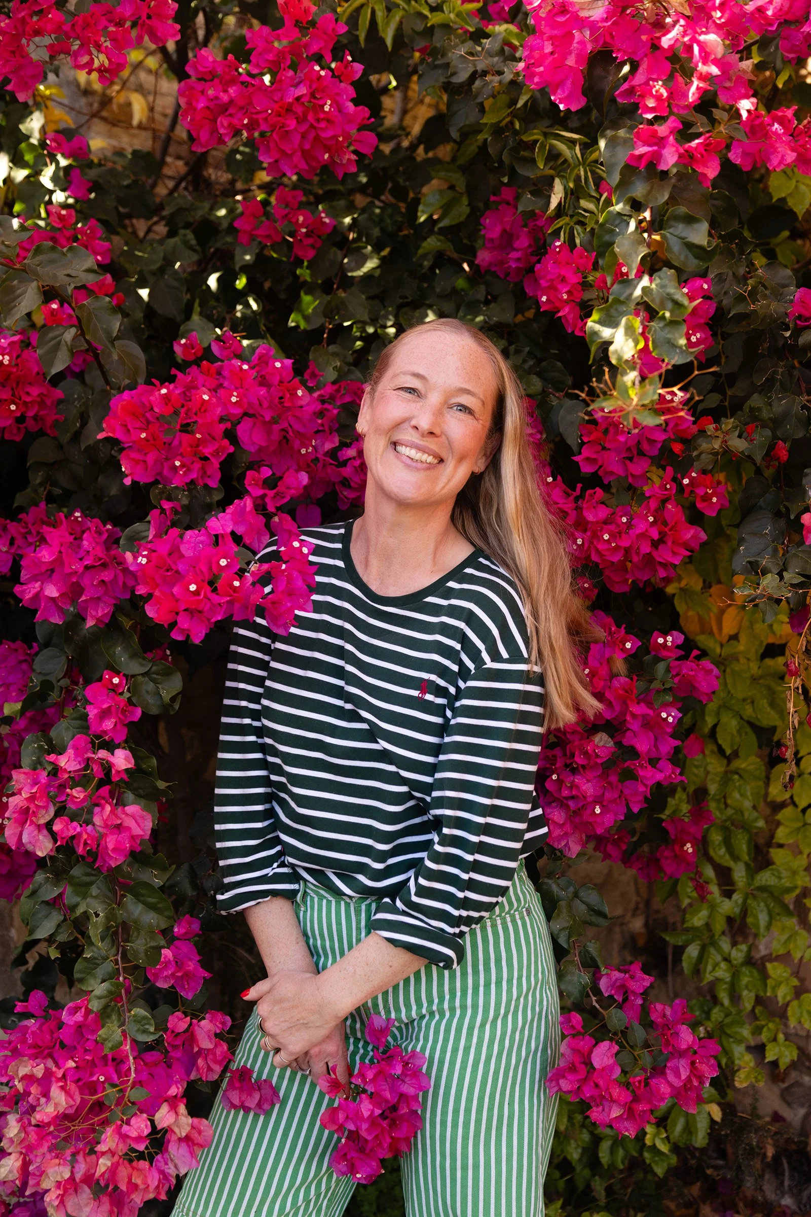 A woman with long blonde hair smiling among vibrant pink bougainvillea flowers, wearing a black and white striped shirt and green striped pants.