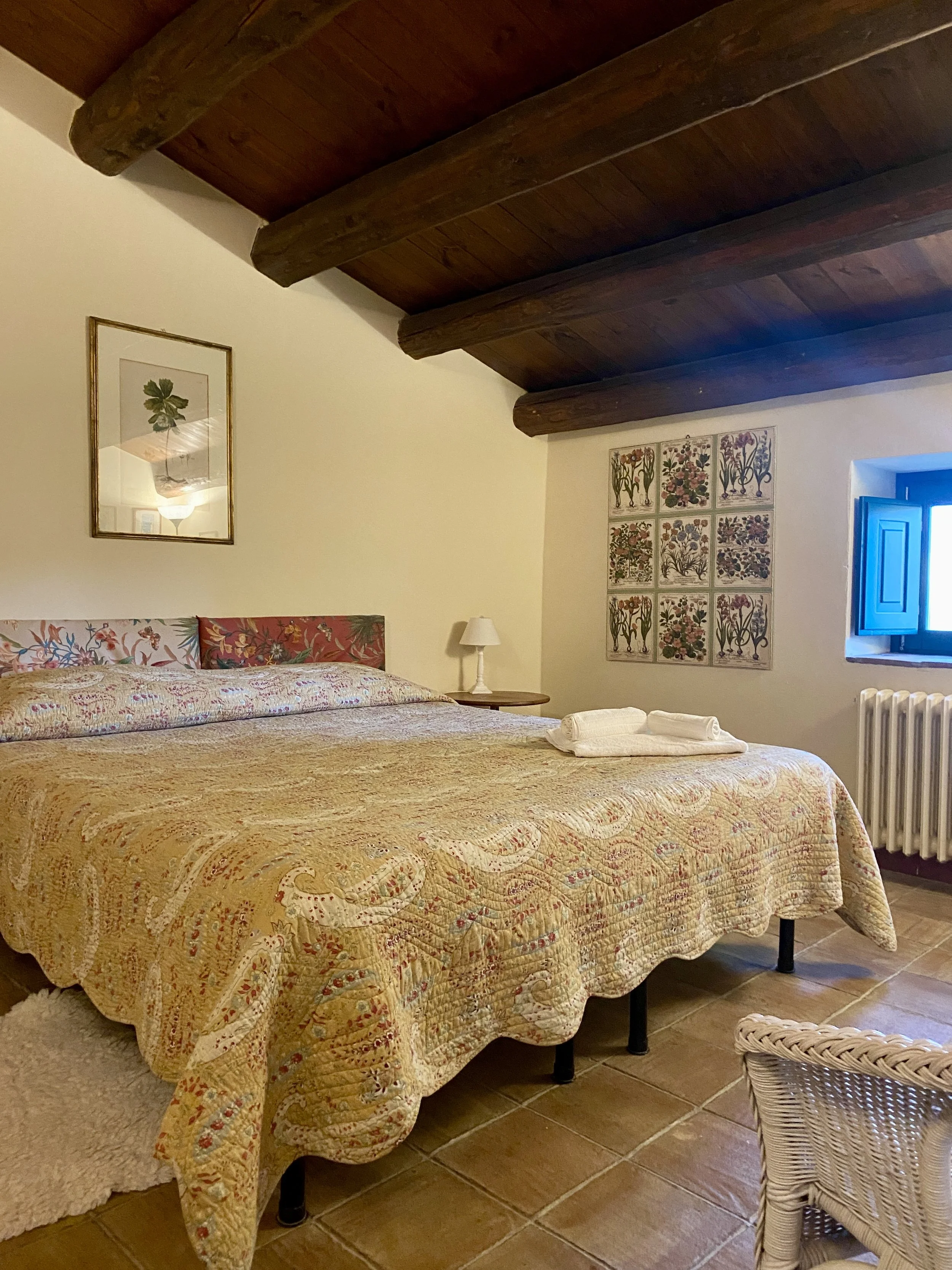 Cozy bedroom with a large bed covered in a patterned yellow bedspread, wooden ceiling beams, window with blue shutters, and artwork on the walls.