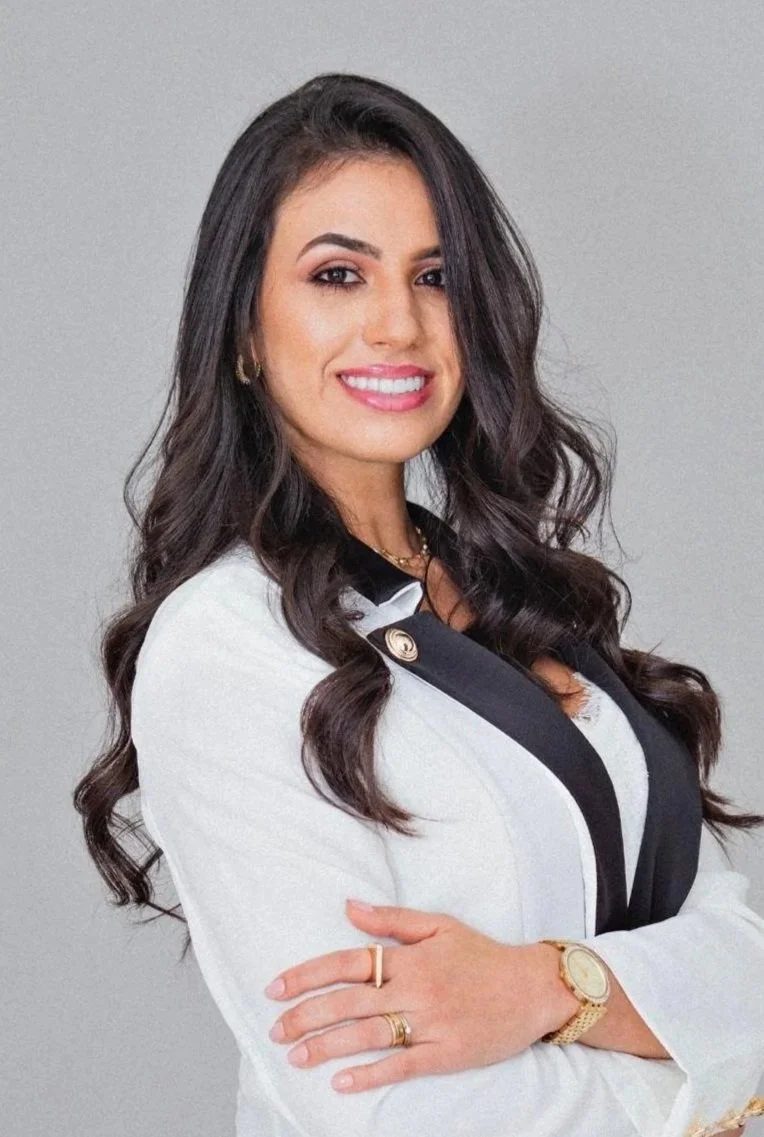 A woman with long dark hair, smiling, wearing a white blazer with black trim, gold jewelry, and a watch, against a light gray background.
