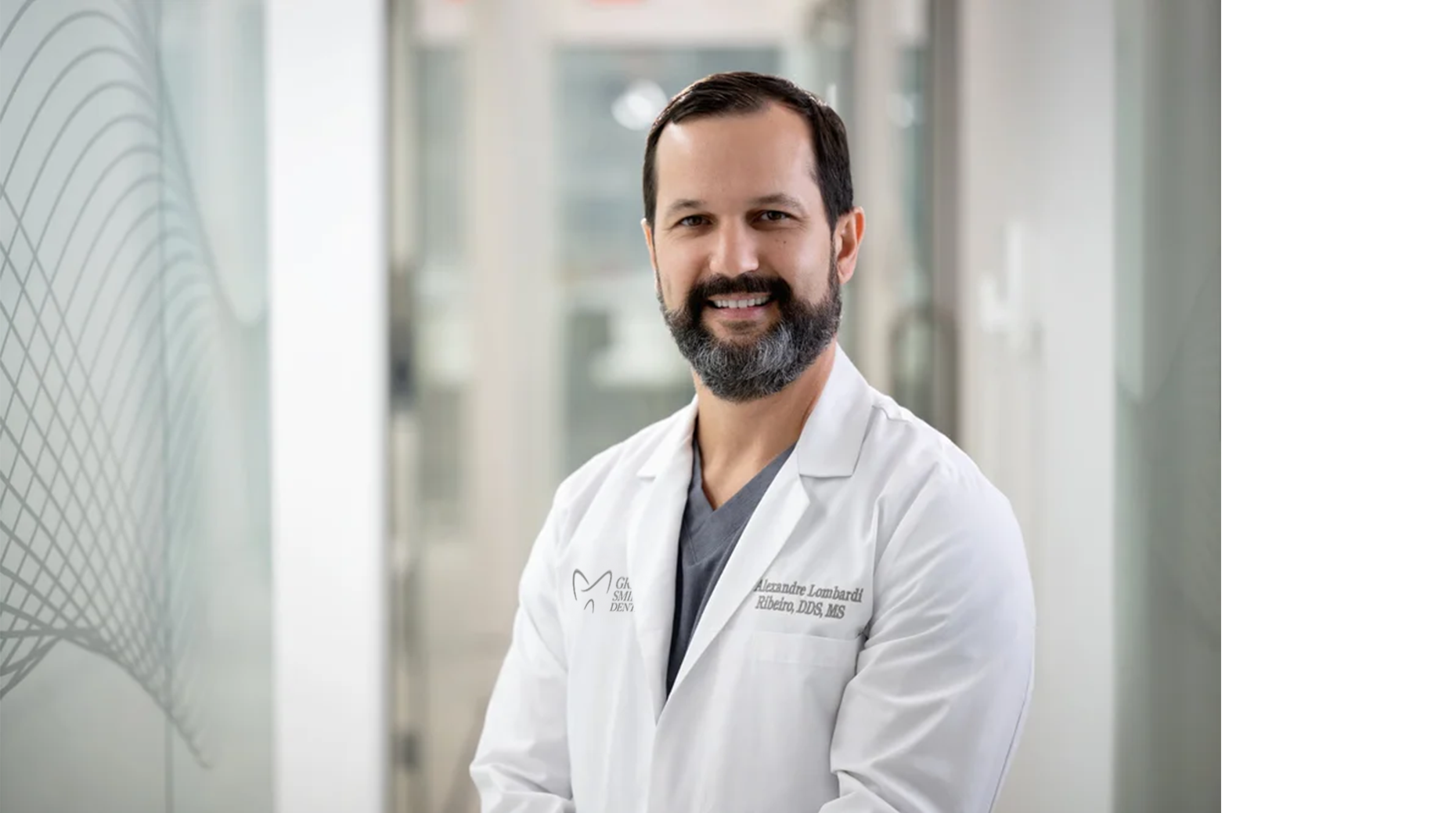 A smiling male doctor with a beard in a white coat, standing in a bright hospital corridor.