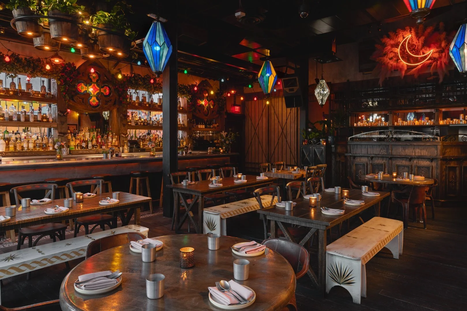 Cozy, dimly lit restaurant interior with wooden tables, benches, a bar area, colorful hanging lights, and decorative wall art.