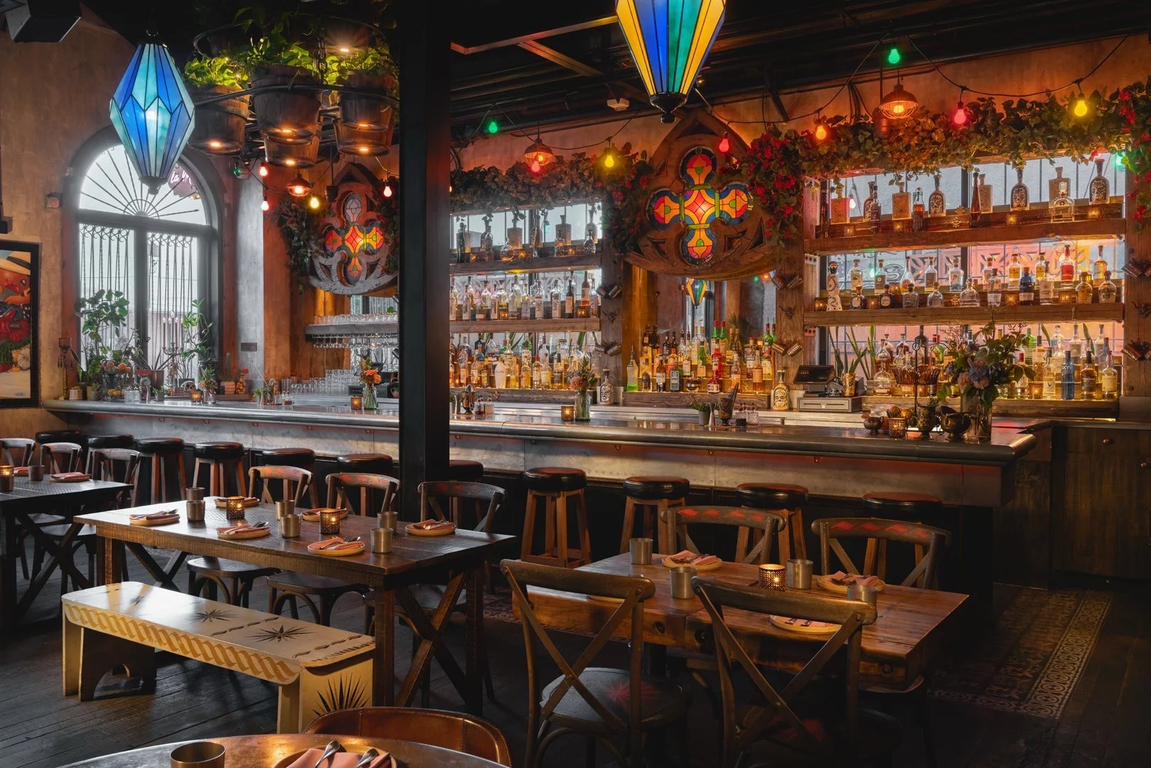 Interior of a cozy, dimly-lit bar with colorful hanging lanterns, a well-stocked alcohol display, wooden tables and chairs, rustic decor, and a warm, inviting atmosphere.