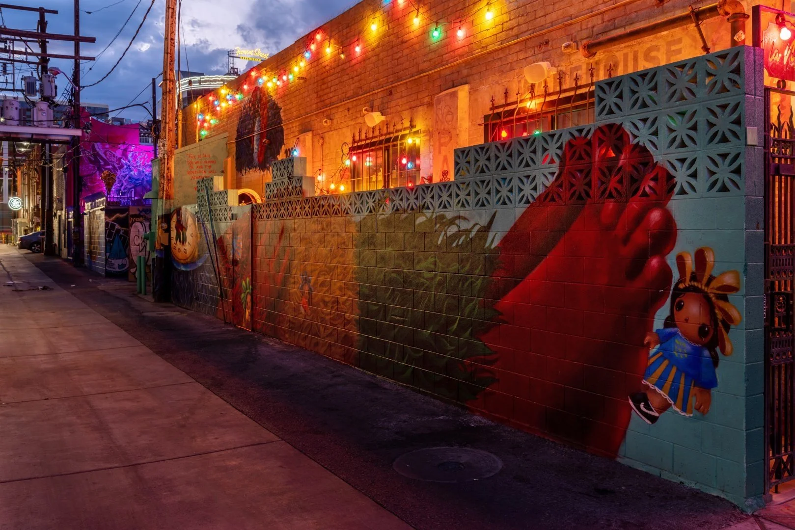 Colorful urban alleyway at night with vibrant graffiti murals on brick walls, featuring playful characters and designs, illuminated by hanging string lights.