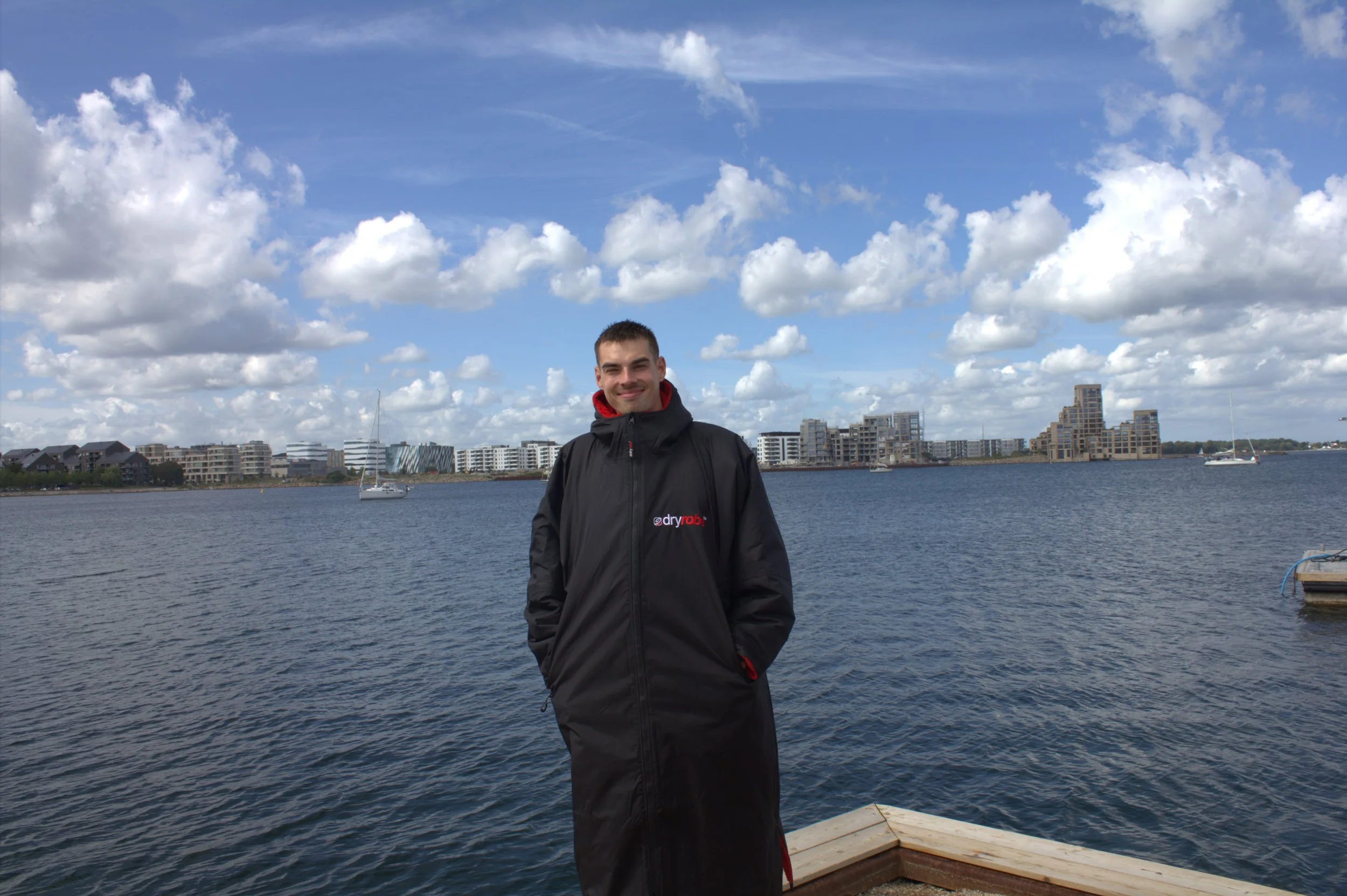 En ung mand står på en mol, iført en sort jakke med logo, foran en waterside med både og moderne bygninger i baggrunden under en skyet himmel.