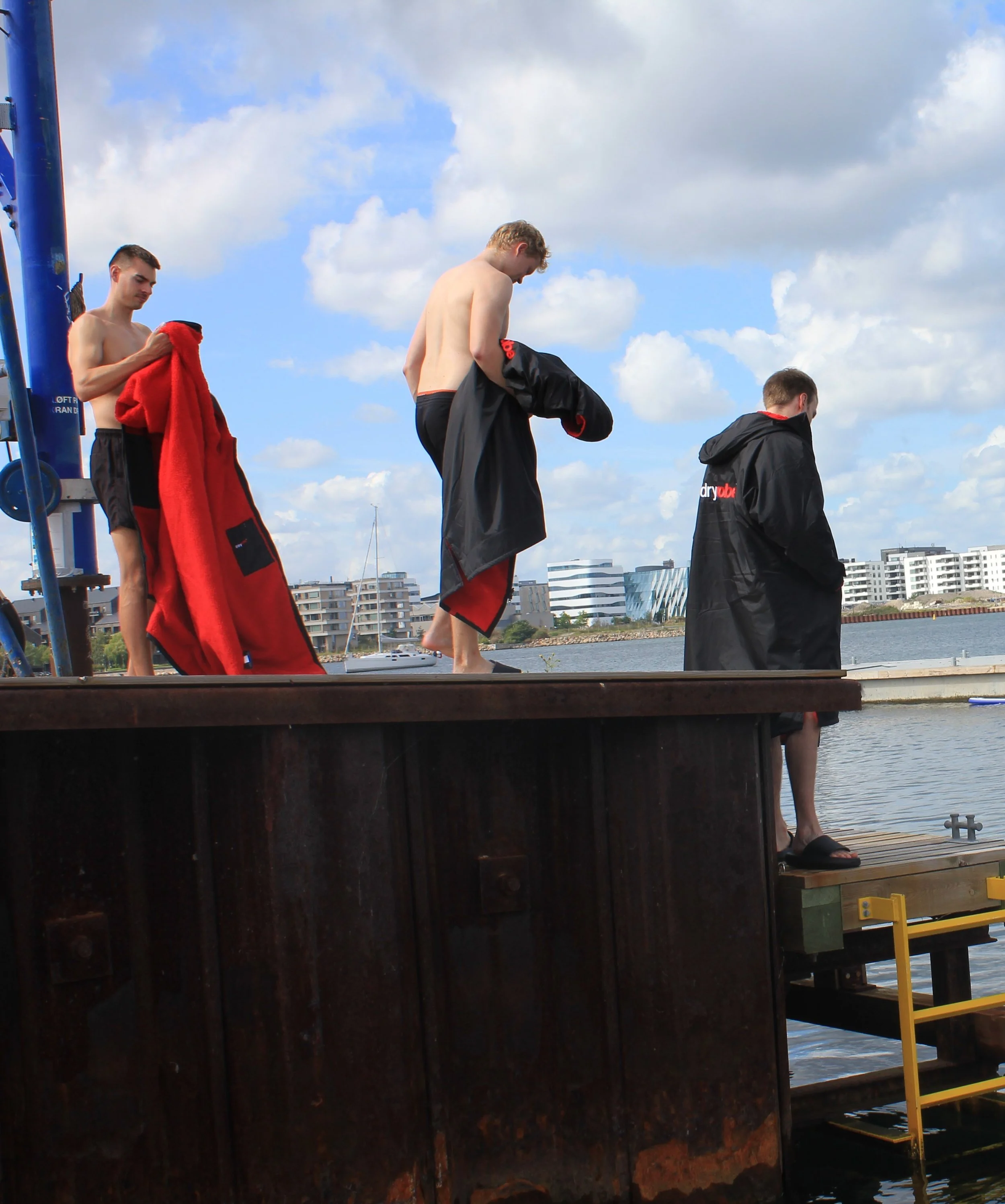 Tre unge mænd er på kajen, nogle er ved at tage badetøj på efter svømning, en kigger ned i sit tøj, og en anden holder tøjet. Baggrunden viser bygninger og en blå himmel med nogle skyer.