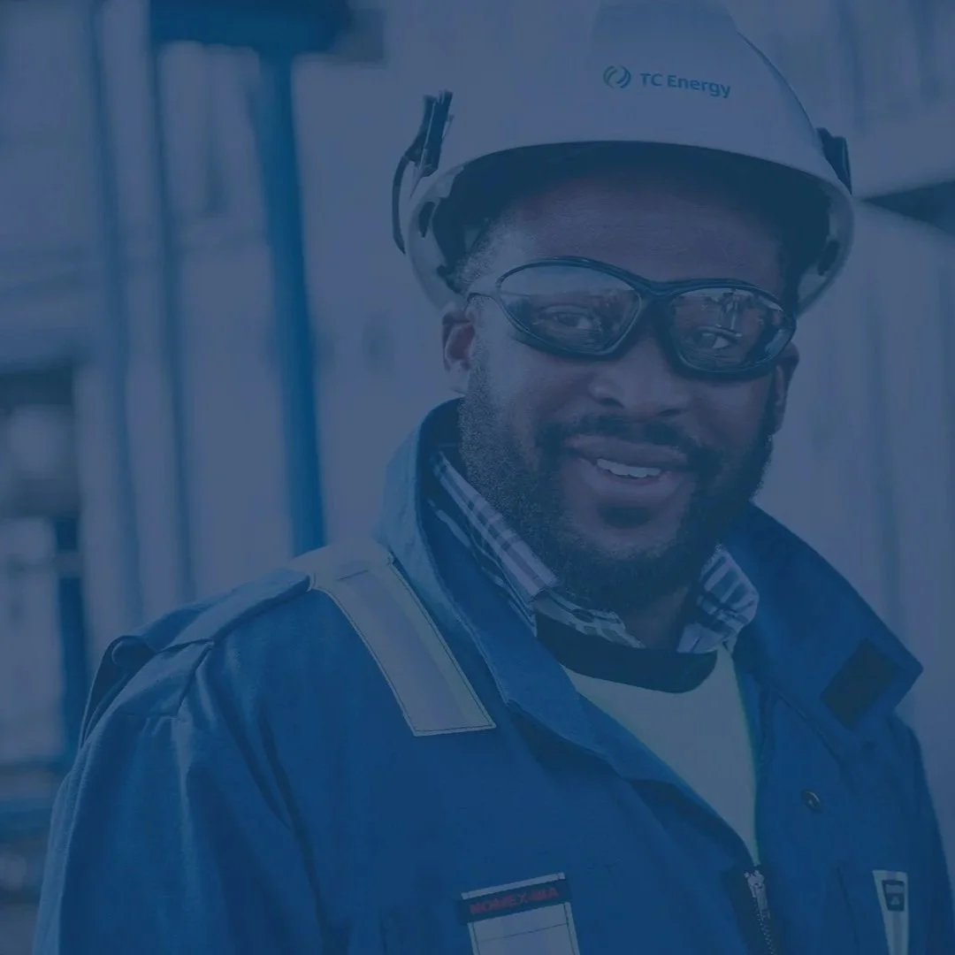 A man wearing safety goggles, a helmet with TC Energy logo, and a blue jacket with a Nomex badge, standing outdoors near a building.