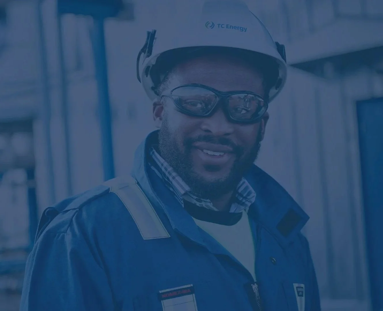 A man wearing safety glasses, a helmet labeled 'TC Energy,' and a blue safety jacket, smiling at a construction site.