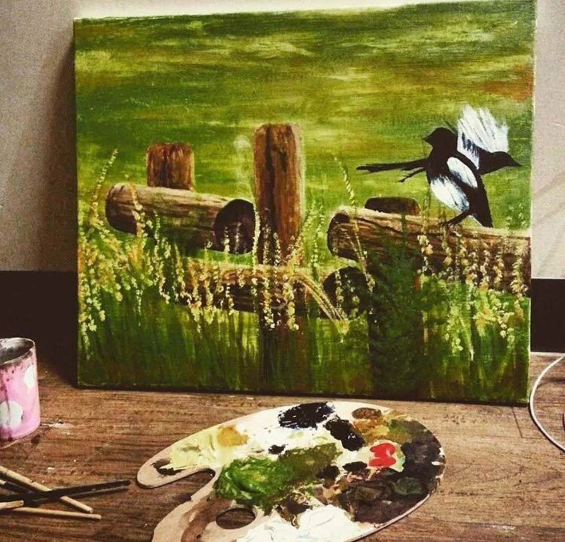 A painting of a rustic scene with fallen logs and a bird perched on one log, set against a green background of grass and foliage. Paint supplies are visible in the foreground.