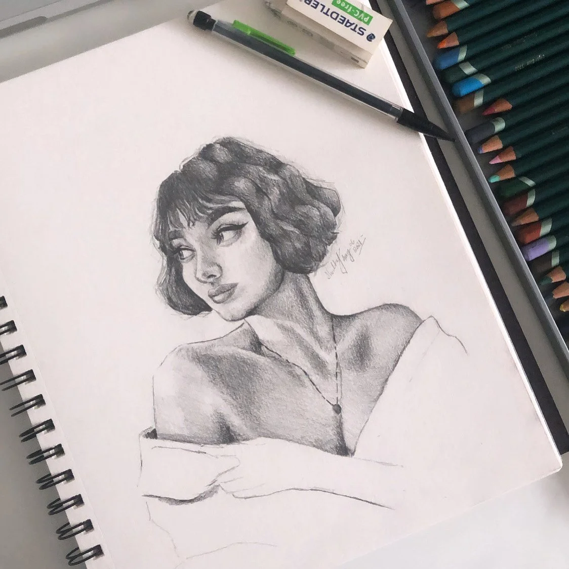 A detailed graphite pencil sketch of a woman with short, wavy hair and a contemplative expression. The sketch includes shading and highlights, and the woman is wearing an off-the-shoulder top and a necklace. Art supplies such as colored pencils, a ma