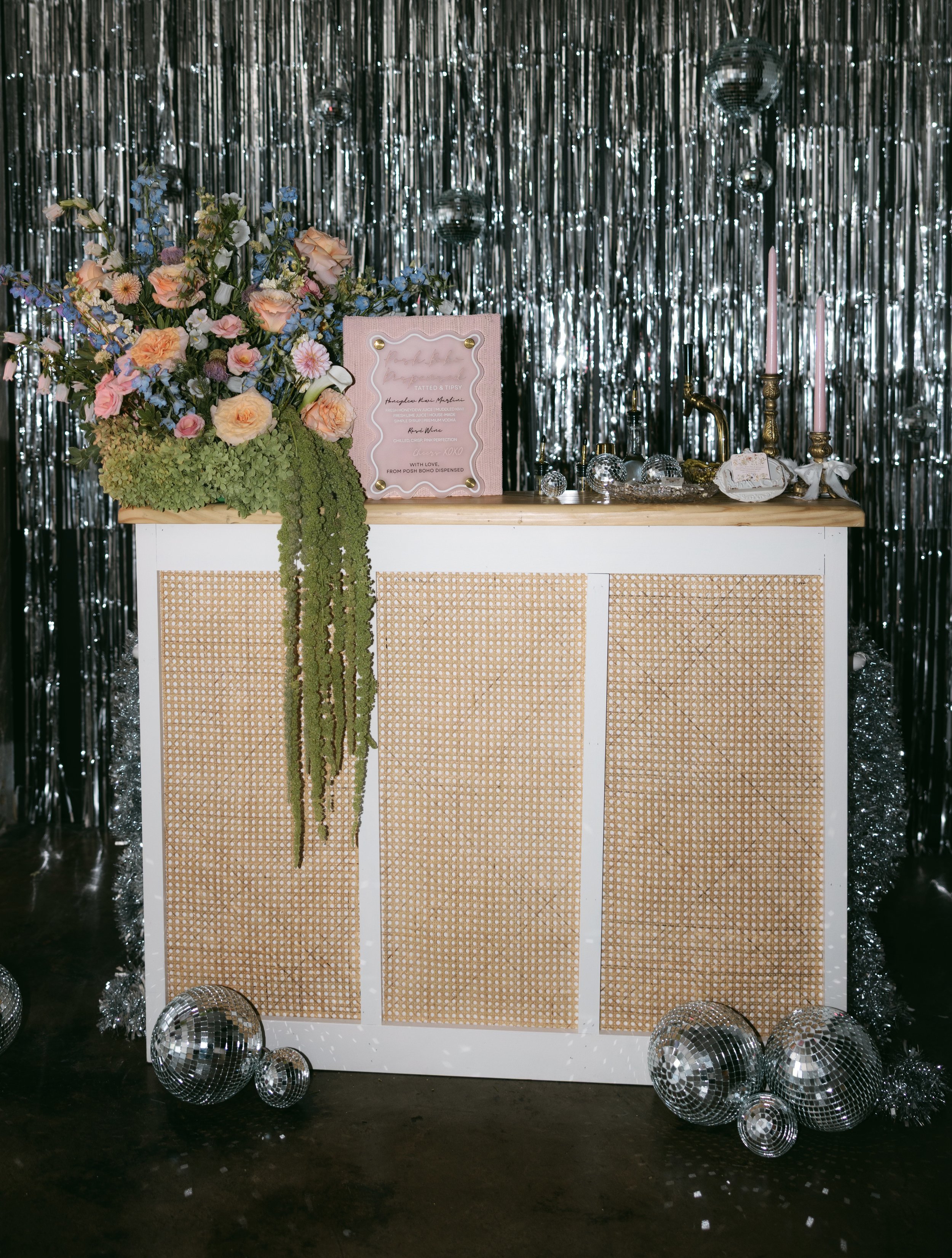 Decorated bar with floral arrangement, pink framed sign, candles, disco balls, and metallic fringe backdrop.