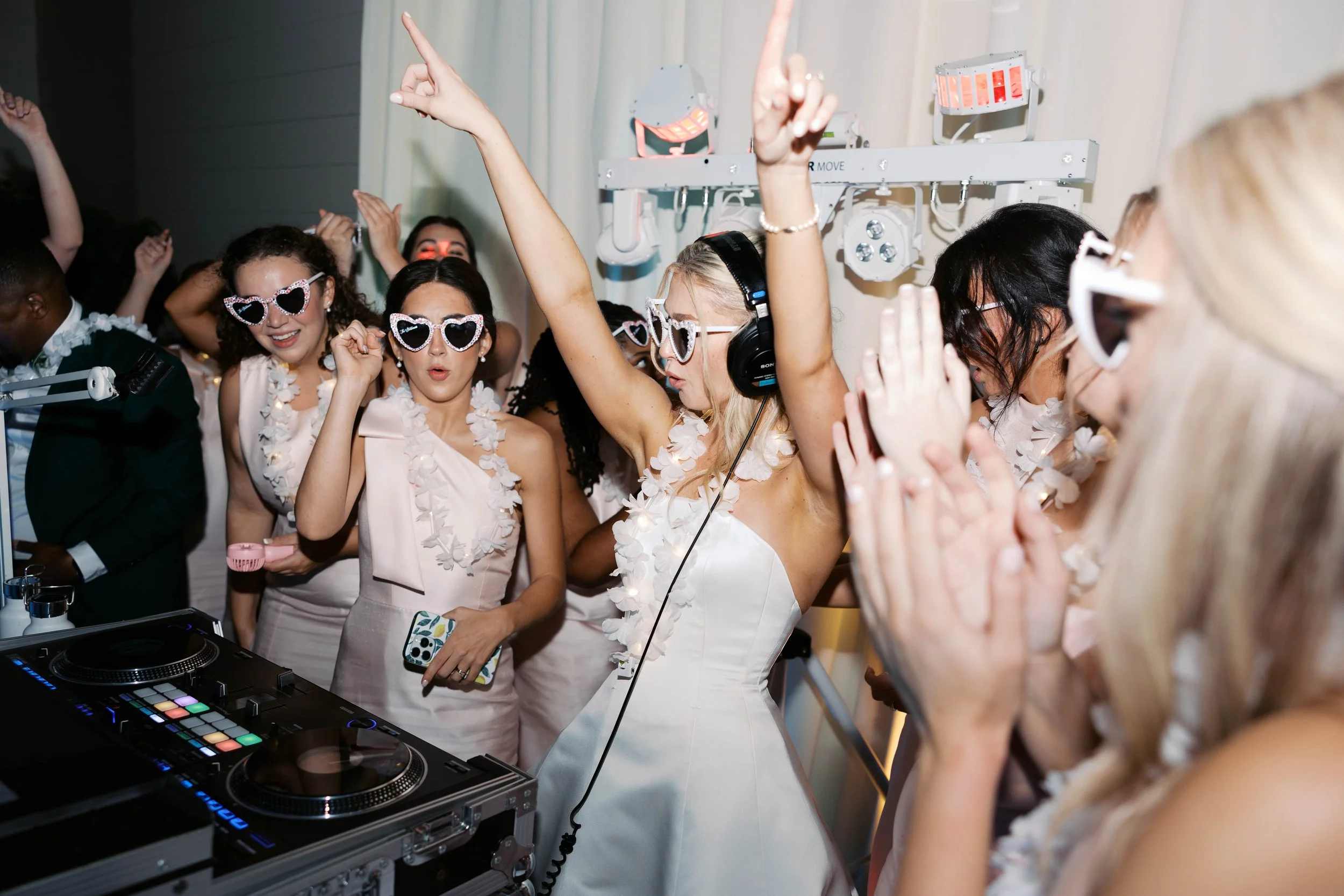 Group of women dancing at a wedding reception, wearing white dresses and sunglasses, with a DJ playing music in the foreground.