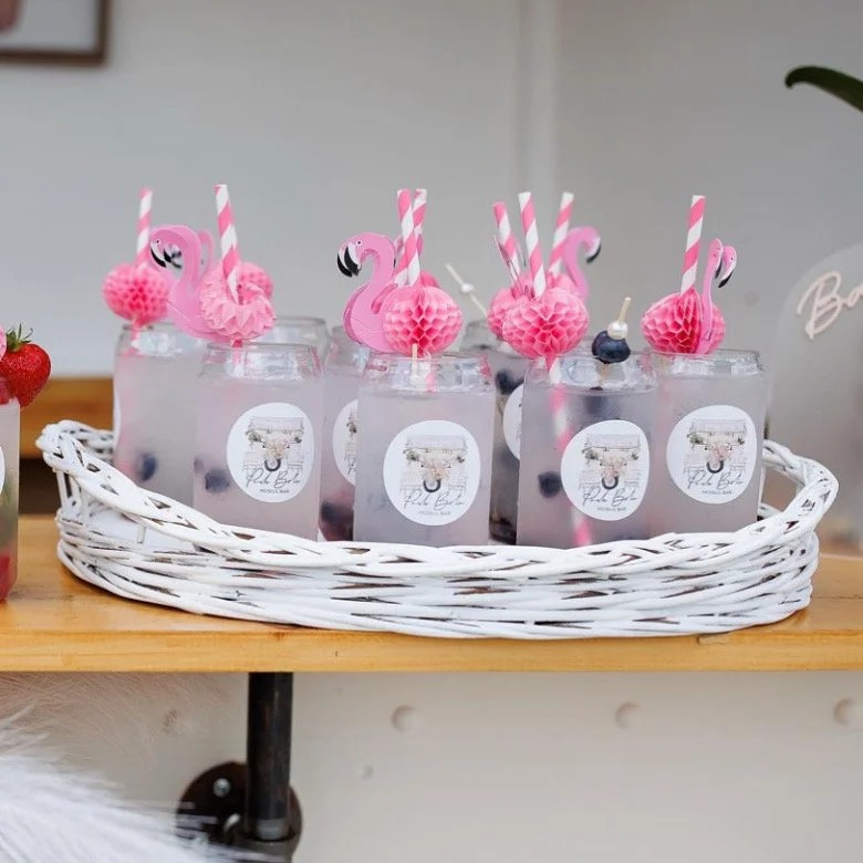 Glass jars with pink flamingo and honeycomb decorations, pink striped straws, and fruit inside, placed in a white wicker tray.
