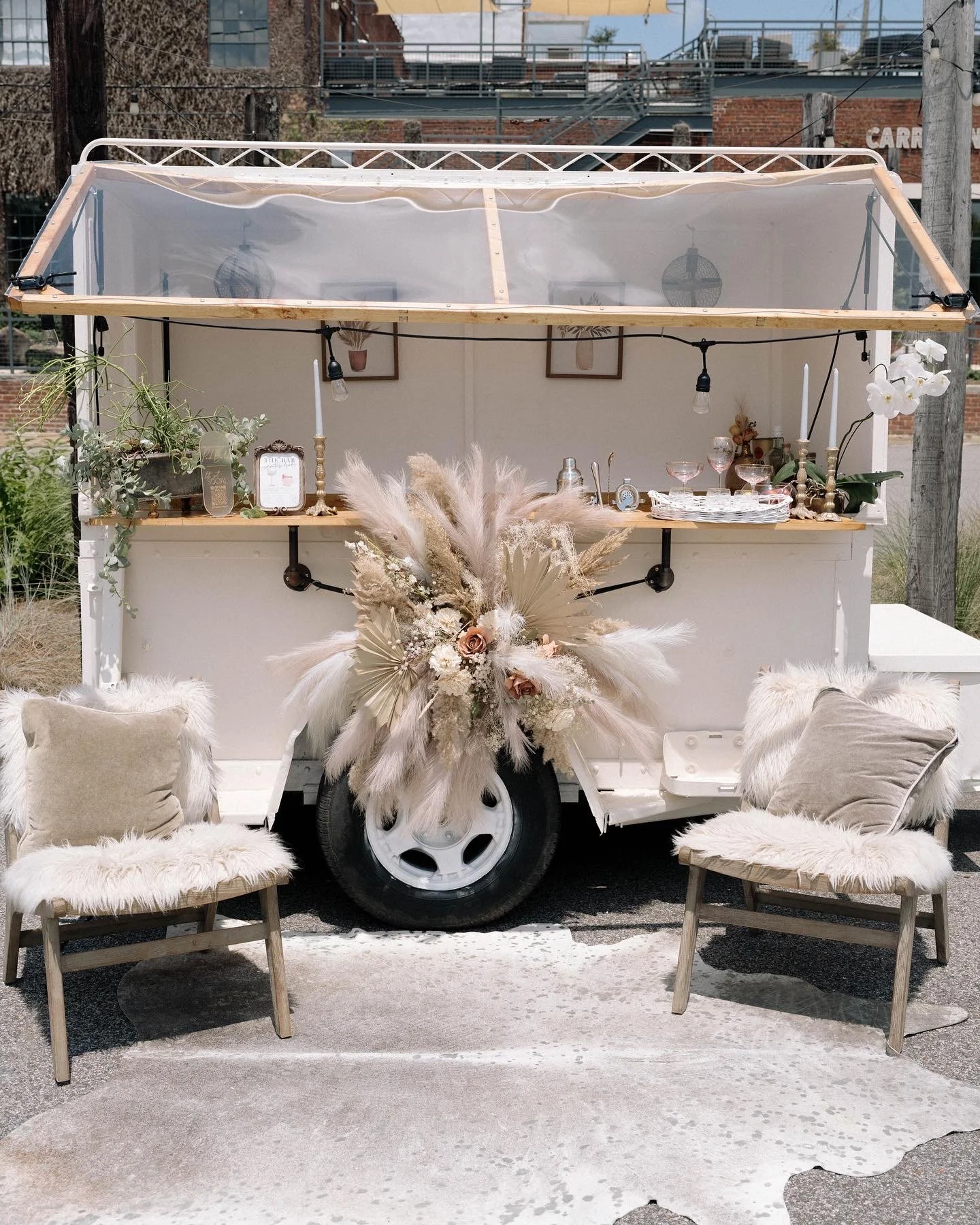 A mobile bar trailer decorated with neutral-colored pampas grass and flowers, with chairs covered in faux fur blankets in front.