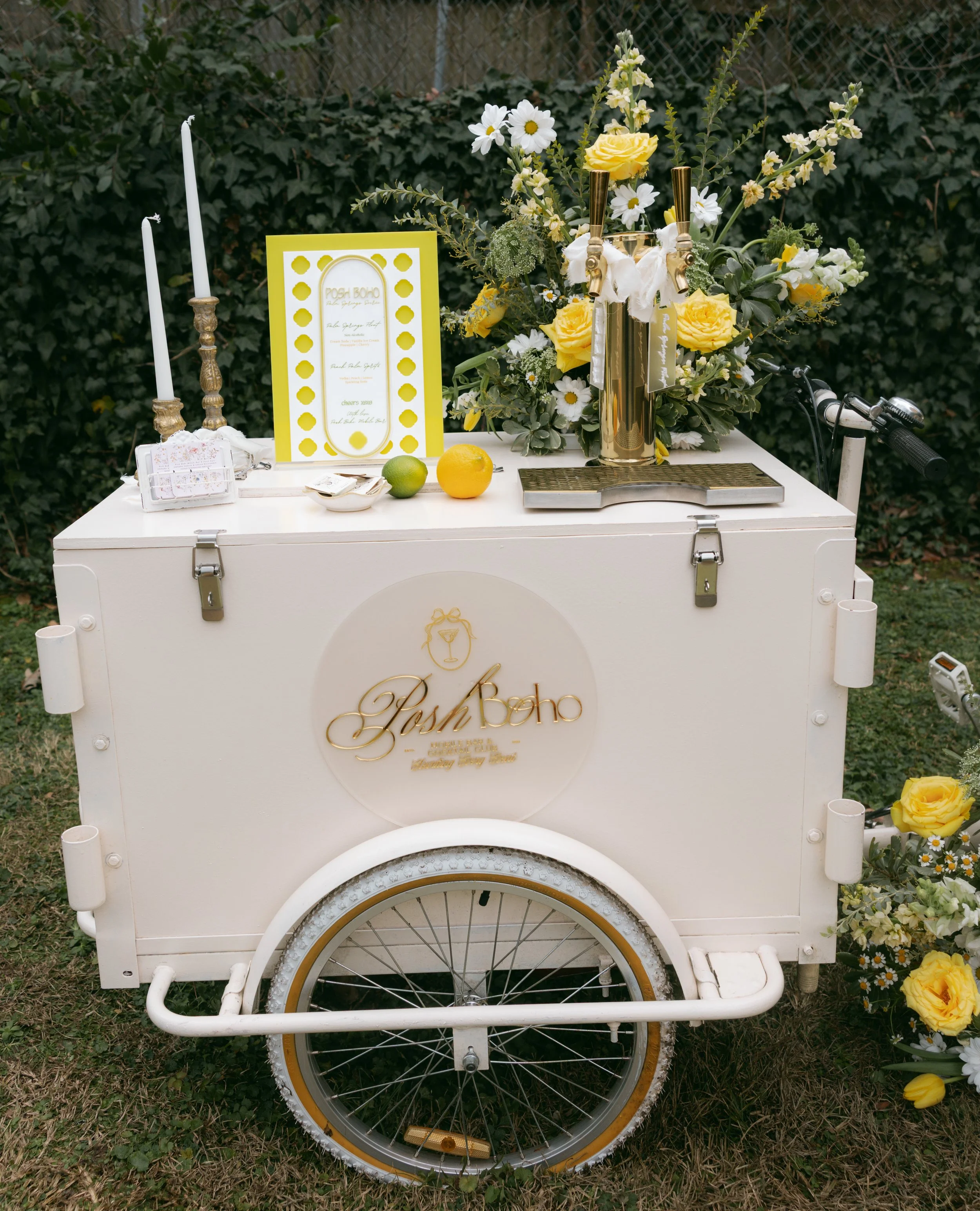 White mobile bar cart decorated with yellow and white flowers, candles, and a menu, set outdoors on grass with greenery in the background.