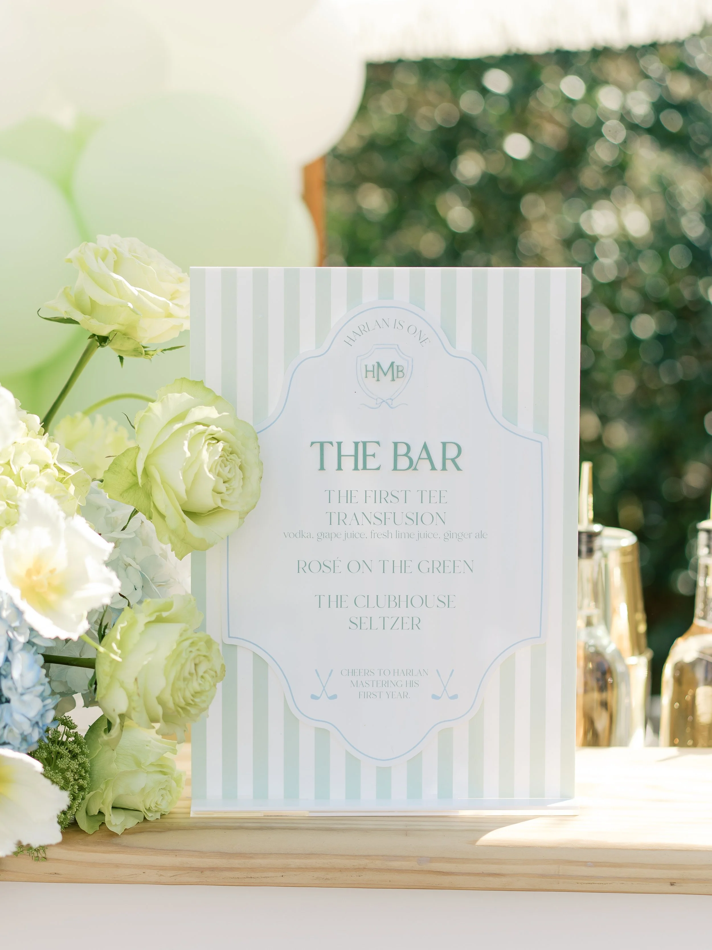 A sign for a wedding bar menu, decorated with white and light green flowers, featuring drinks like vodka, grape juice, lime juice, ginger ale, and seltzers, with a background of balloons and foliage.