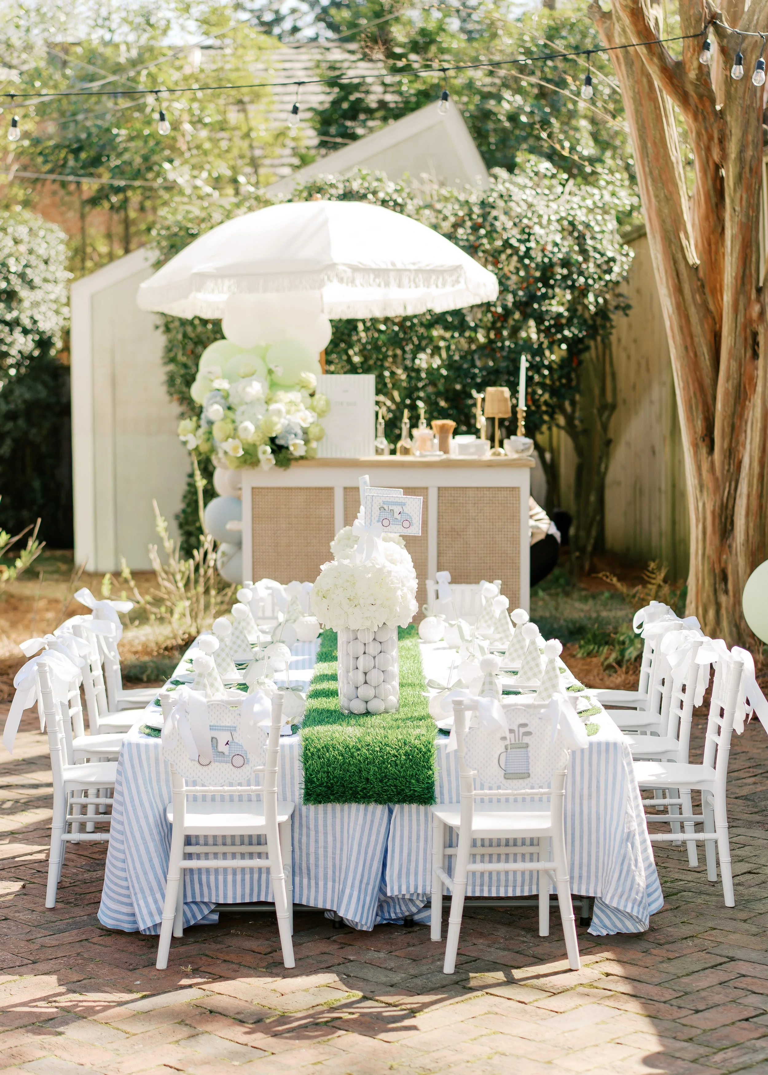 Outdoor celebration setup with a long table covered in a blue and white striped tablecloth, decorated with a green grassy runner and white decor. Small white chairs with bows face the table. In the background, there is a wooden bar with a white umbrella, flowers, and balloons, under string lights and surrounded by trees and bushes.