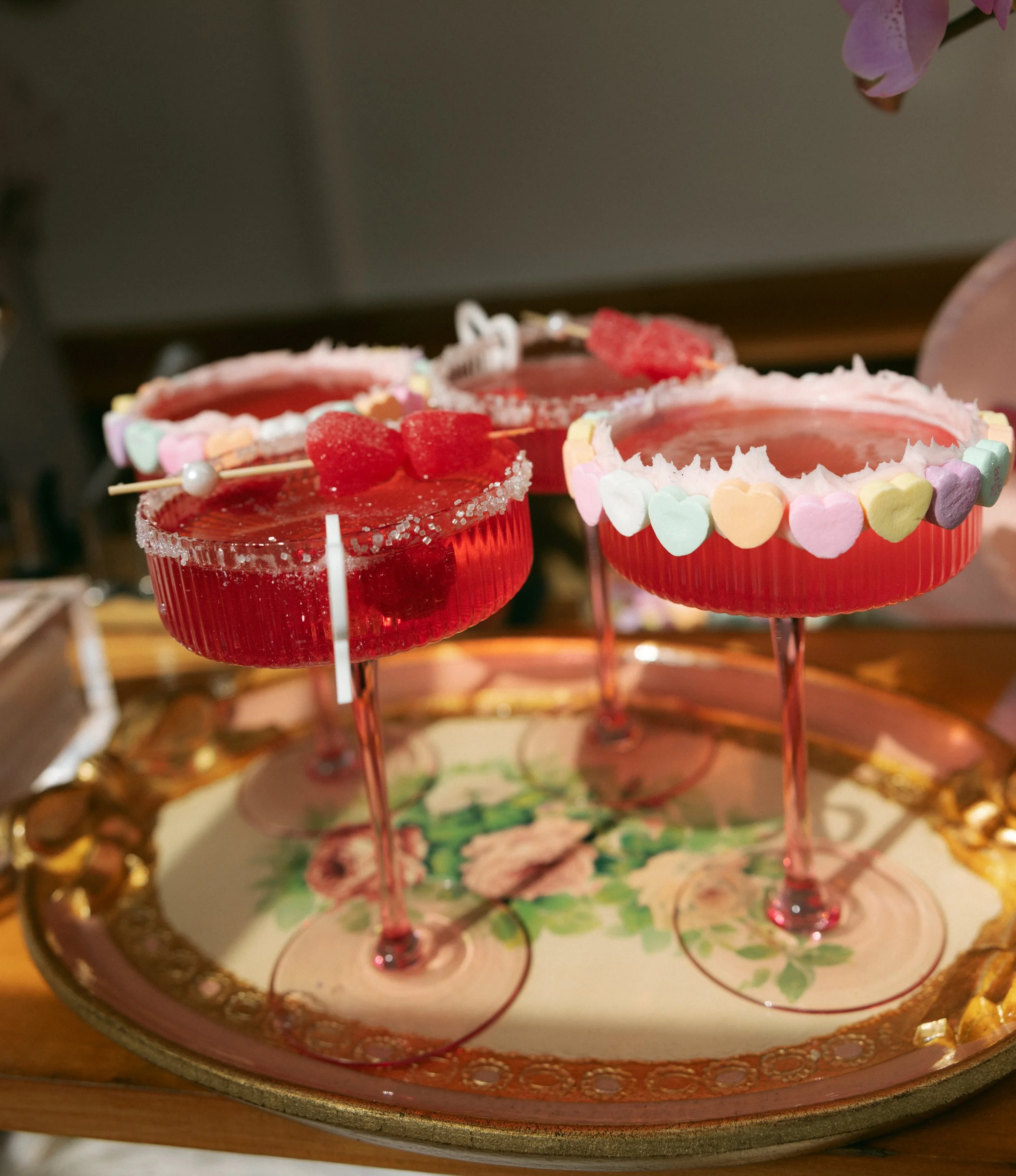 Three pink cocktails with sugar rims, garnished with gummy bears and lollipops, on a floral serving tray.