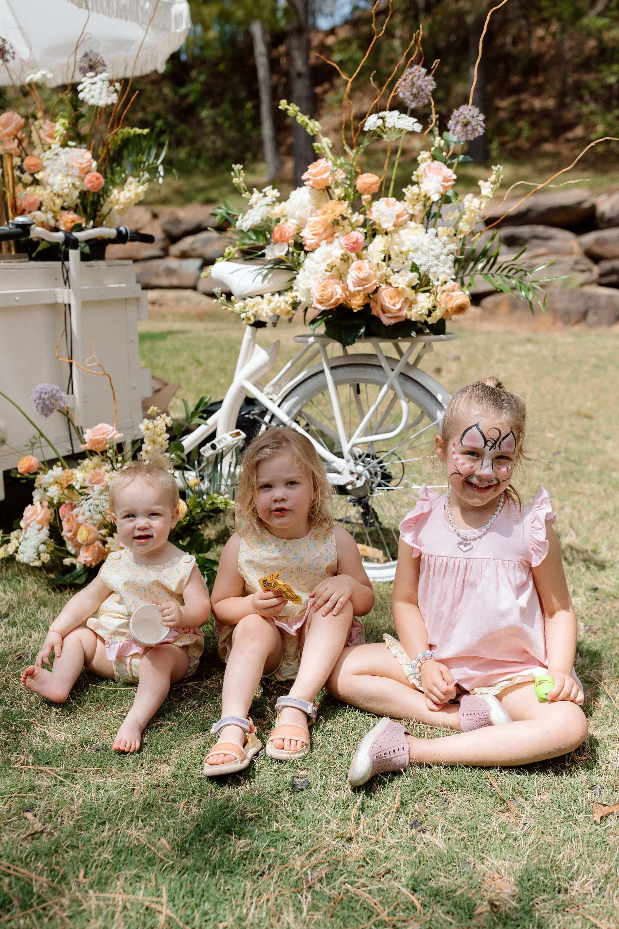 Three young girls sitting on the grass during a celebration, with a decorated white bicycle and floral arrangements in the background.