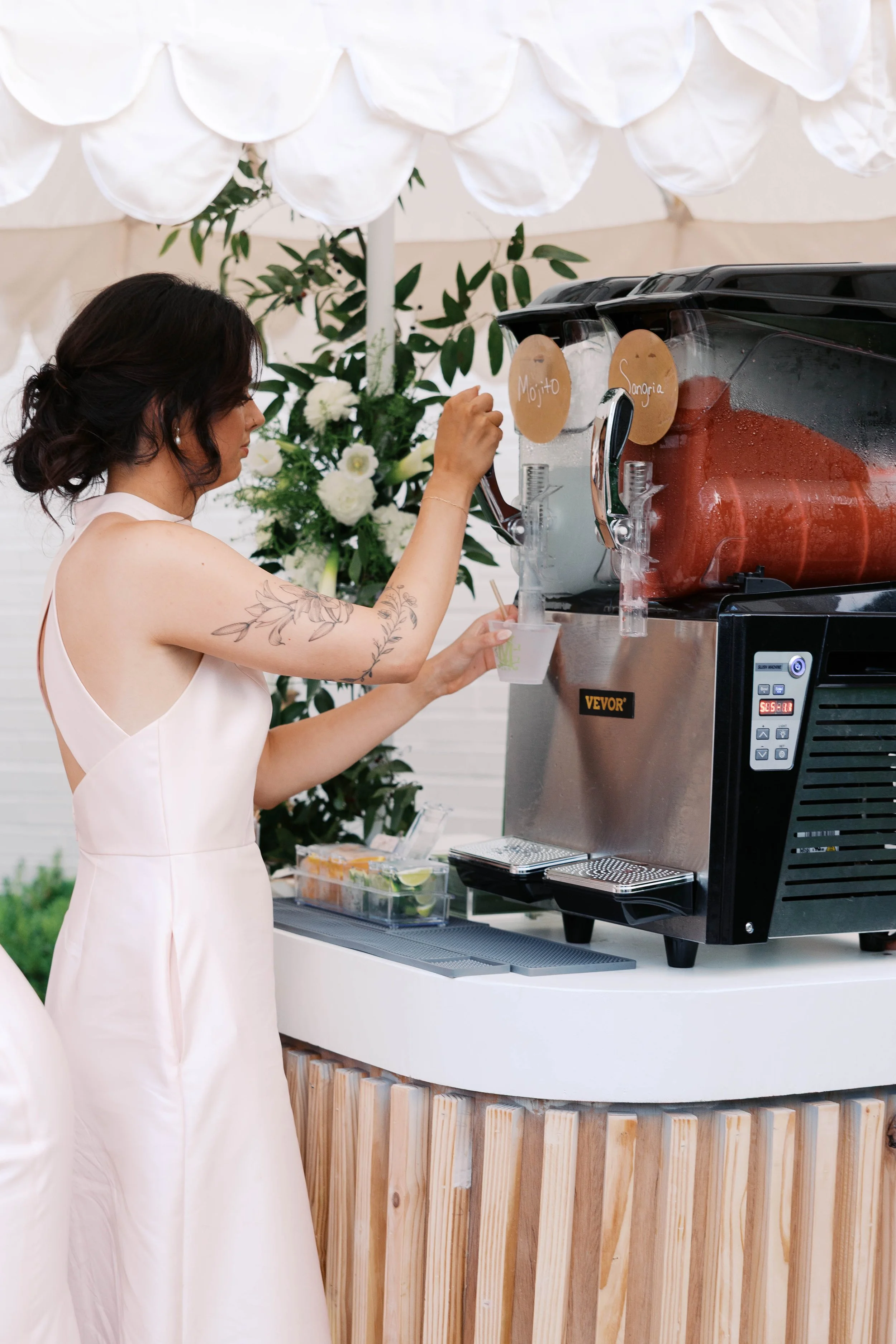 A woman in a white dress pouring drinks from a beverage dispenser, with signs indicating mojito and sangria, at an outdoor event with decorative flowers and a white canopy.