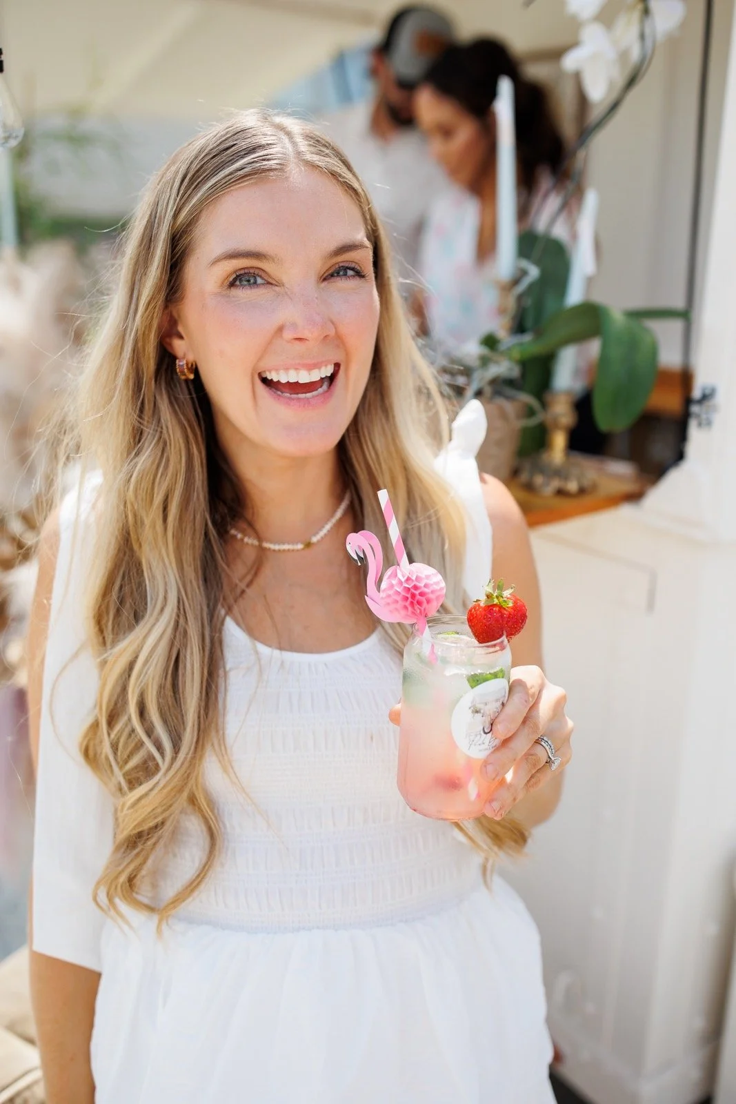 A woman with long blonde hair smiling and holding a pink drink decorated with a strawberry and flamingo-shaped straw at a social gathering.