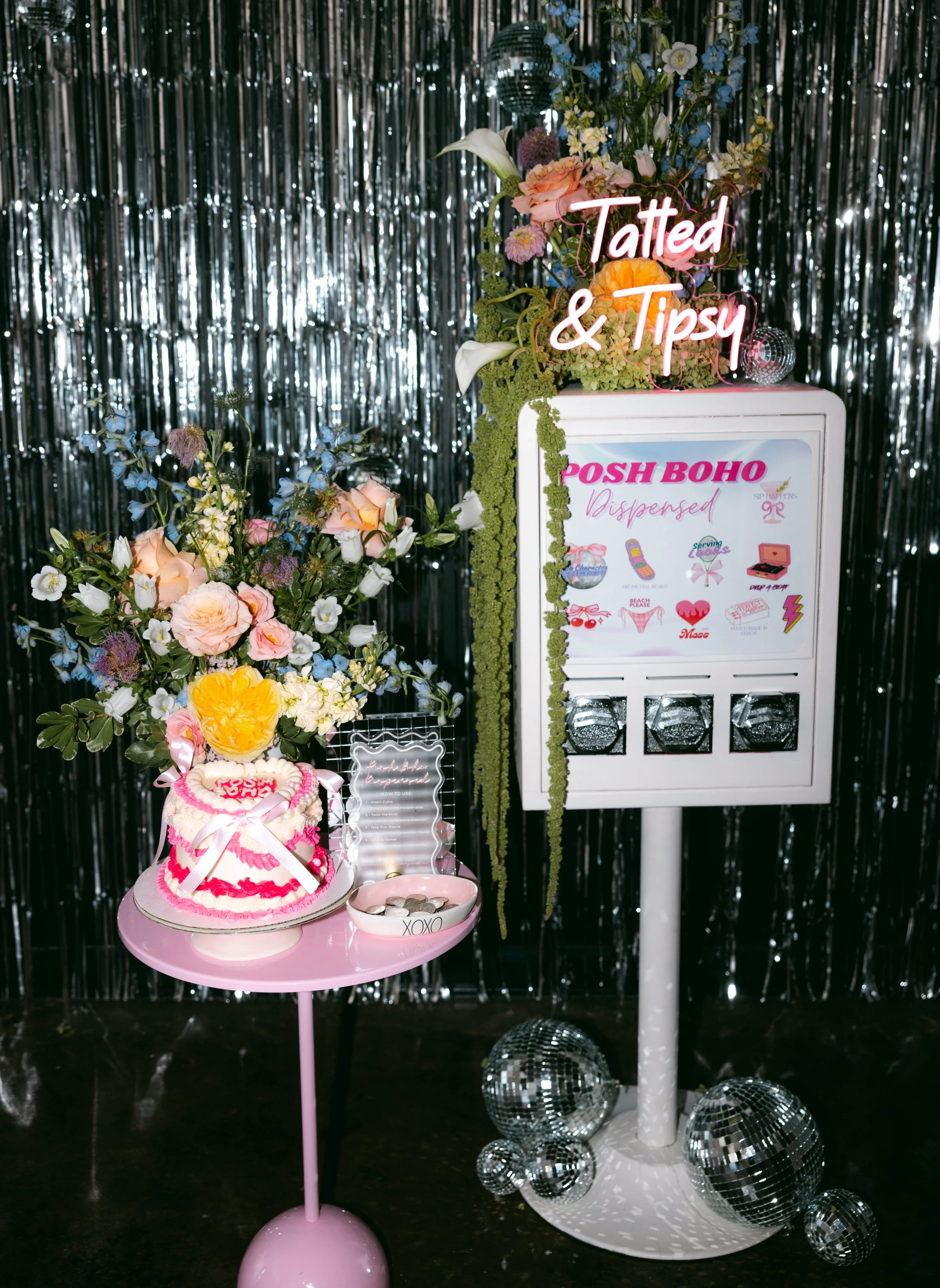 Decorative display featuring a pink cake with white and pink icing, a large flower arrangement, a pink table, a dispenser box labeled 'POSH BOHO Dispersed,' a floral arrangement with a neon sign that says 'Tatted & Tipsy,' and shiny disco balls against a metallic fringe backdrop.