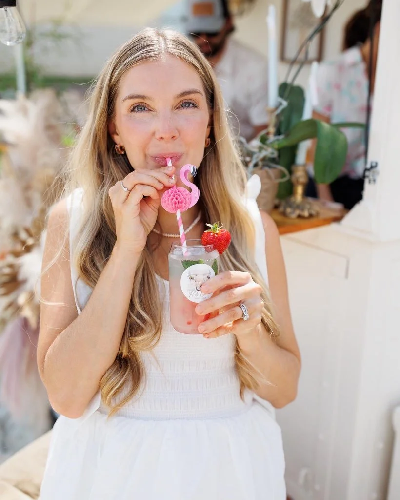A woman with long blonde hair enjoying a beverage with a pink flamingo straw and a strawberry garnish, outdoors with blurred background.