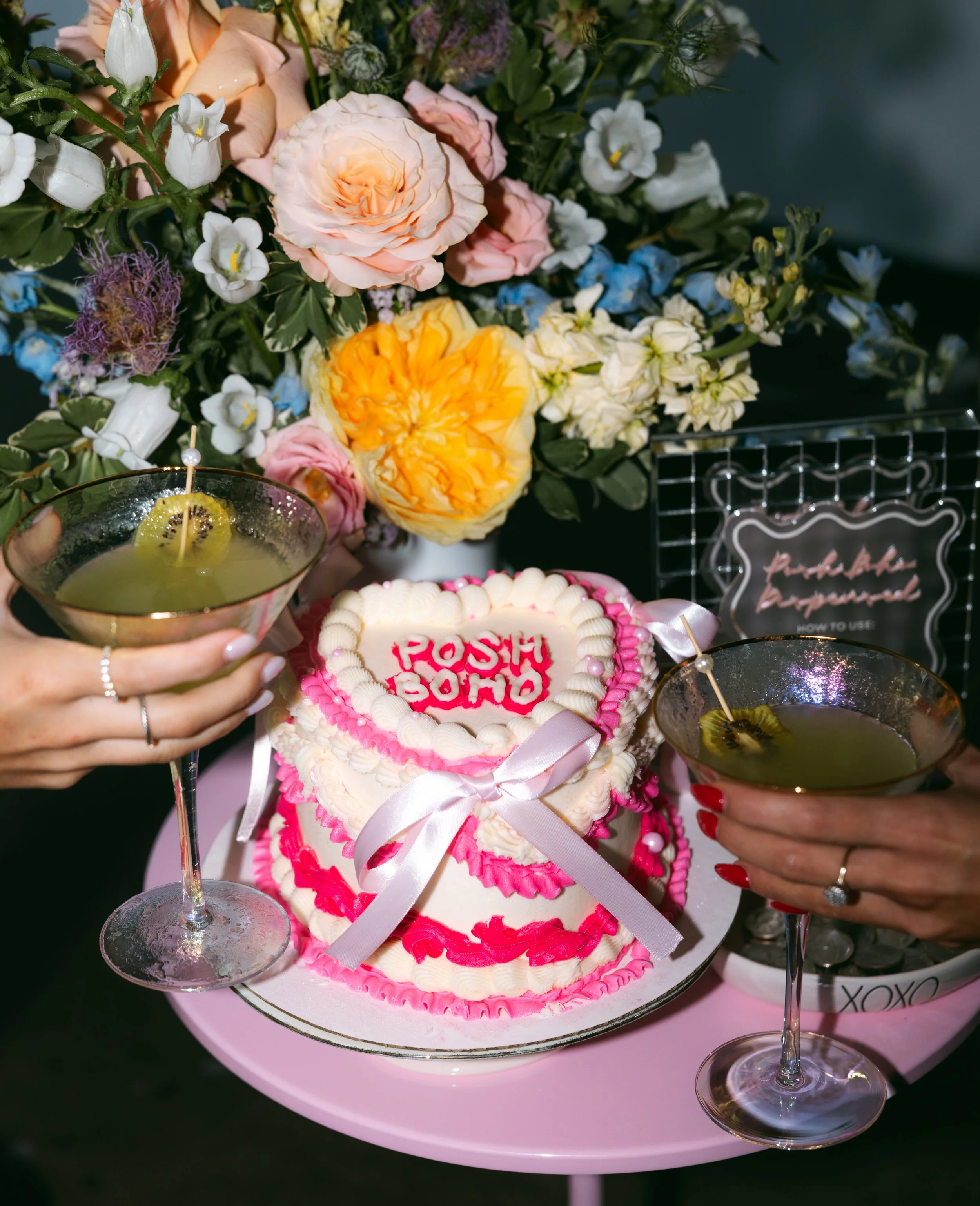 Two women holding martini glasses with kiwi slices for a celebration in front of a heart-shaped cake decorated with pink and white icing and the words 'Posh Pop'. There is a large bouquet of mixed flowers, a small framed chalkboard sign, and pink tab
