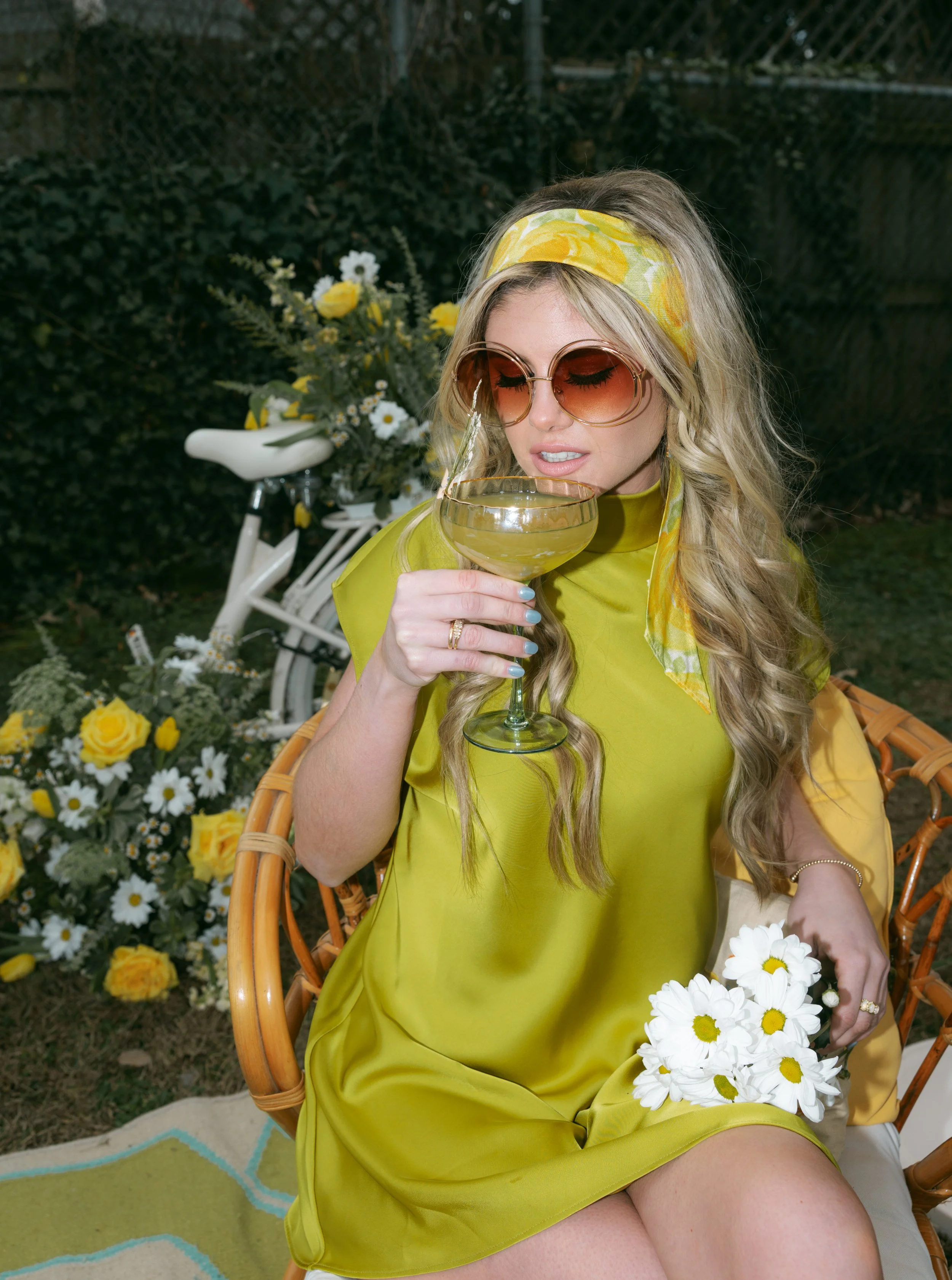 A woman with long wavy blonde hair wearing a bright yellow dress, large sunglasses, and a yellow floral headscarf, sitting outdoors in a garden with flowers and a white bicycle in the background. She is holding a glass of white wine and looking at it.