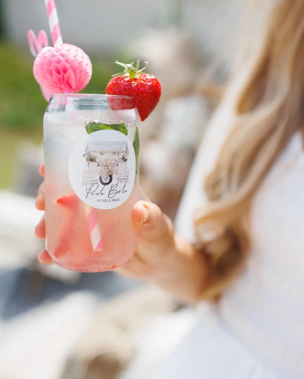 A person holding a glass filled with a pink beverage, garnished with a strawberry and a green mint leaf, with a pink honeycomb-shaped decoration and a pink-striped straw on top.
