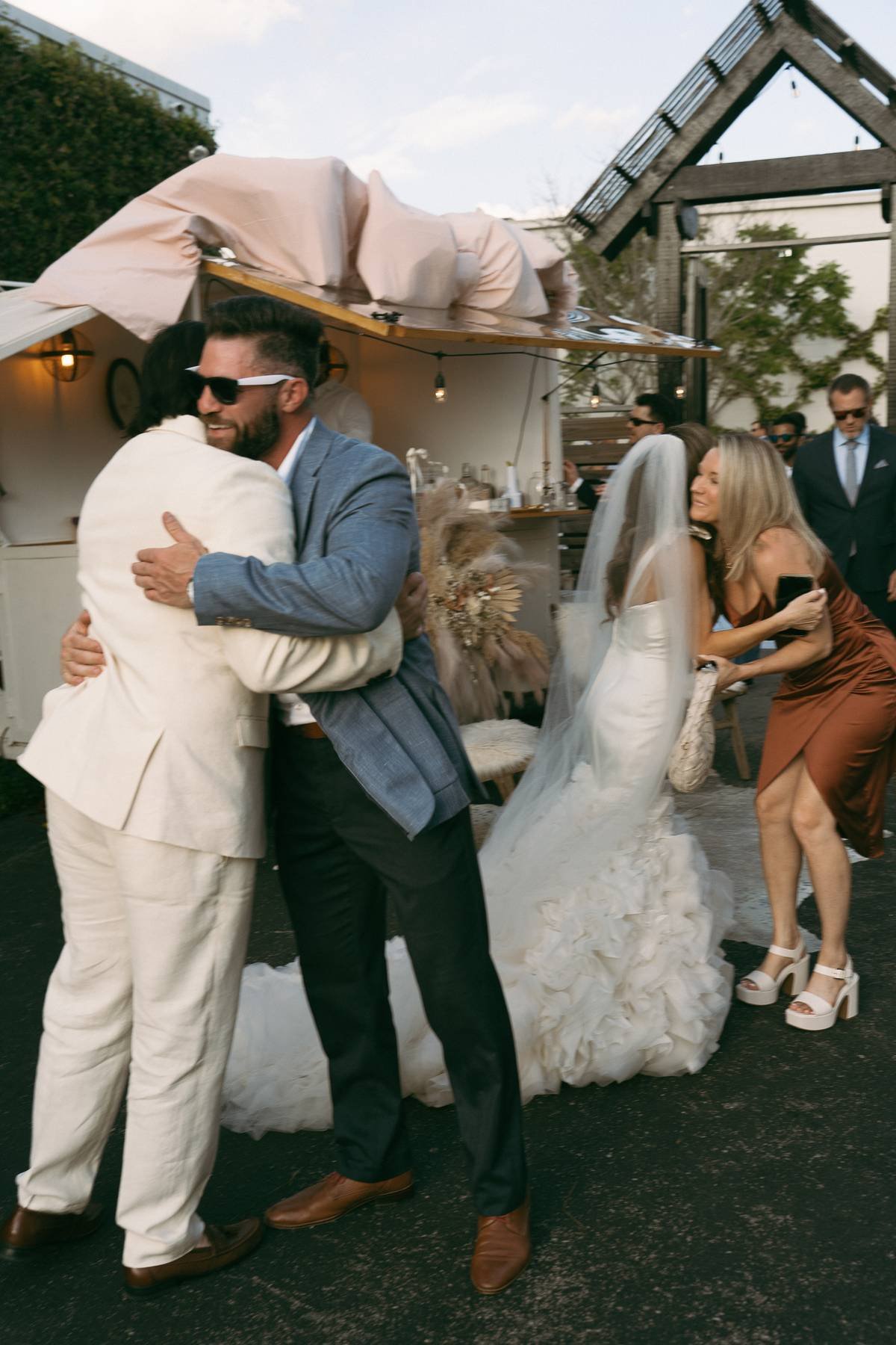 Two men hug warmly in the foreground, one in a cream suit, the other in a blazer and dark pants, at a wedding reception. Behind them, a bride in a white wedding dress with a train and veil is talking to a woman in a rust-colored dress, who is holding