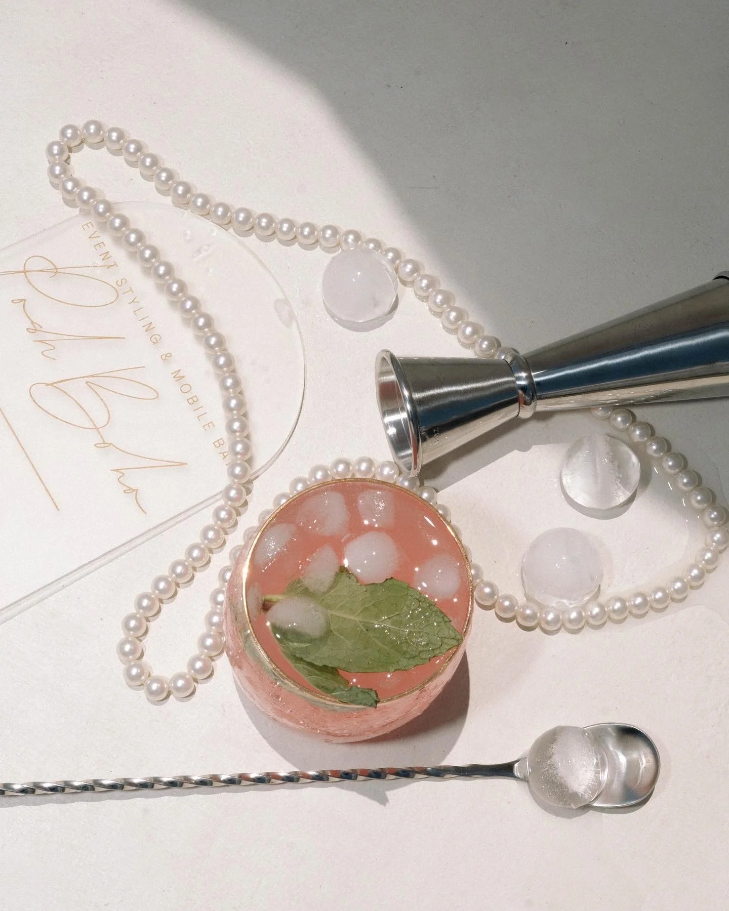 A flat lay of a pearl necklace, a clear glass bowl with a pink drink and ice, a silver cocktail jigger, and a silver cocktail spoon on a white surface, with a blurred business card in the background.