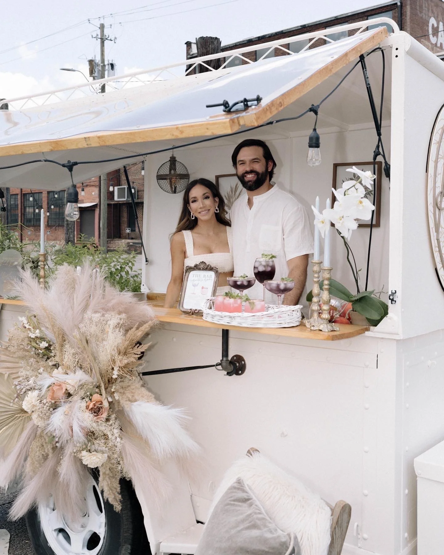 A smiling couple standing inside a white food truck decorated with flowers and candles. The woman is in a white dress, and the man is in a white shirt. There are drinks on the counter, framed menus, and string lights inside the truck.
