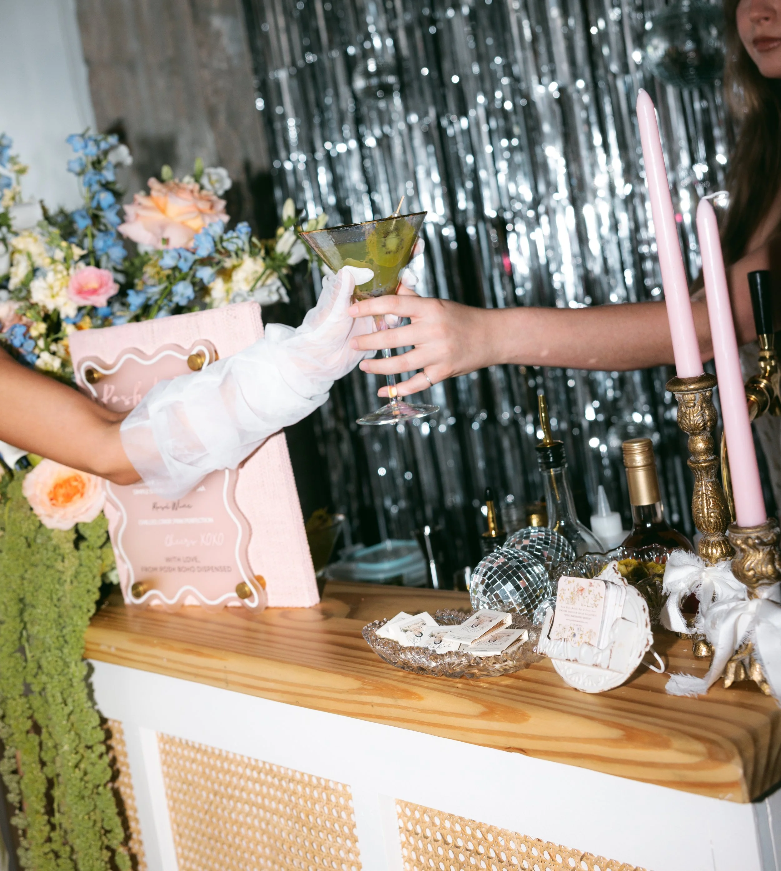 Person in white glove hands a green cocktail in a martini glass to another person at a decorated event table with pink candles, flowers, and a black and silver backdrop.