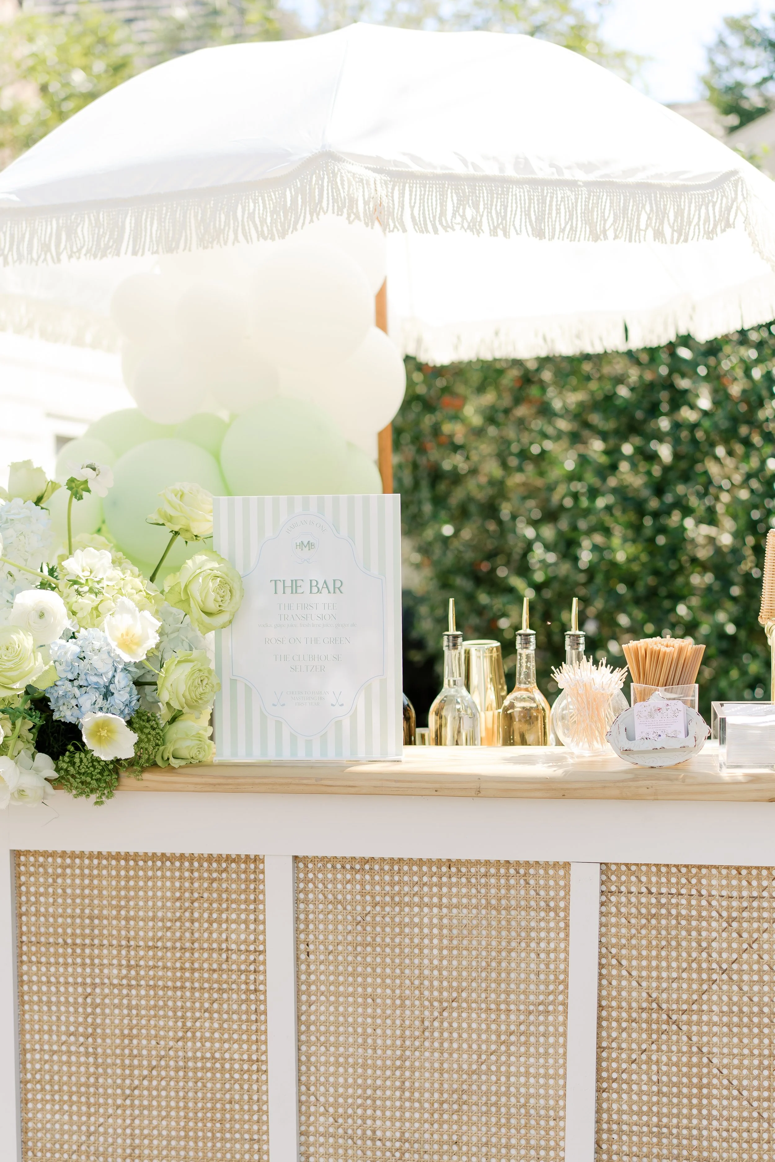 Outdoor bar setup with flowers, balloons, and various bottles under a white parasol.