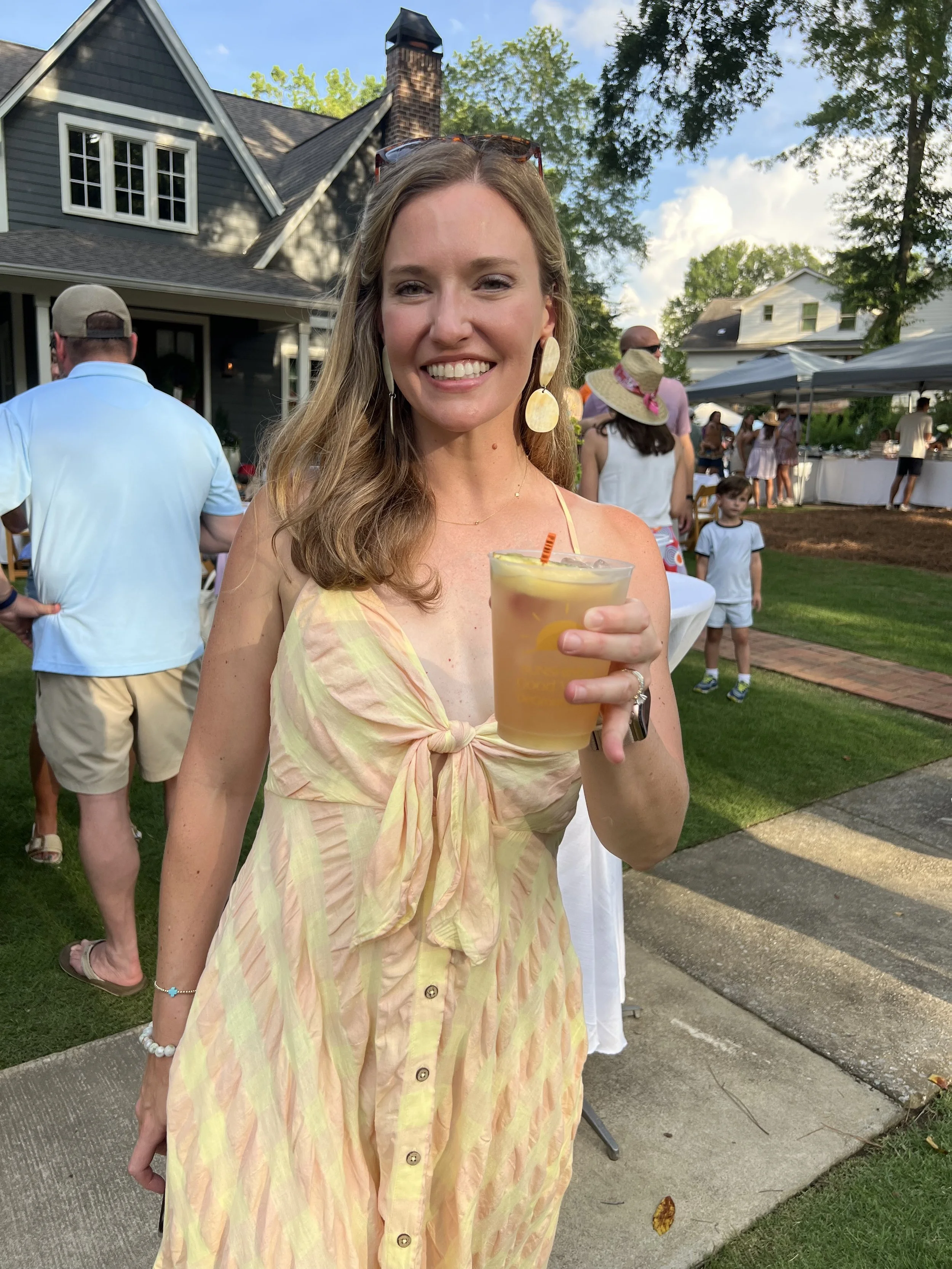 A woman smiling and holding a drink with a lemon slice in an outdoor backyard setting during a gathering or party, with other people and a house in the background.