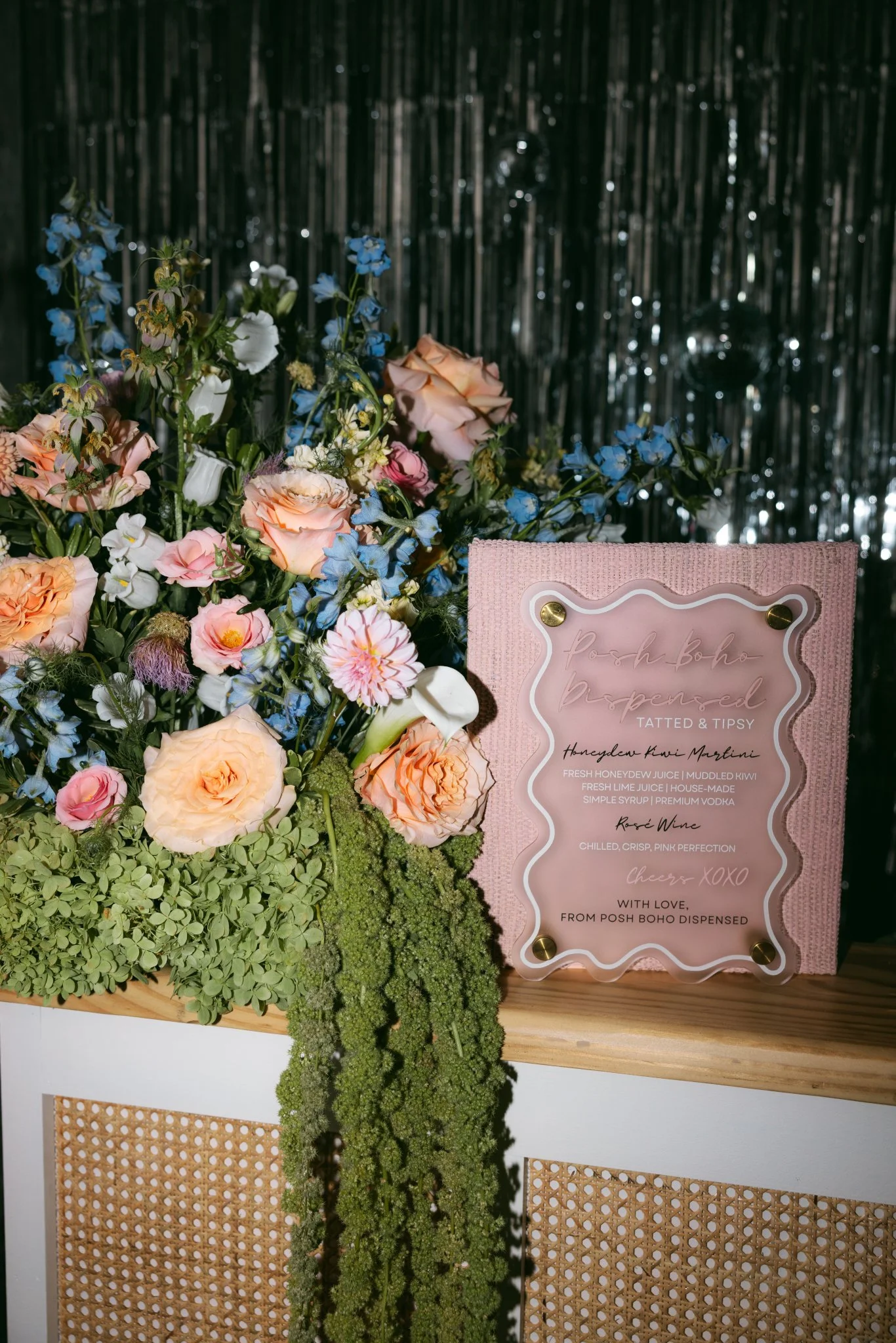 A floral arrangement with peach, pink, and white roses, blue flowers, and green foliage beside a pink, decorative sign on a wooden surface, with a metallic curtain backdrop.