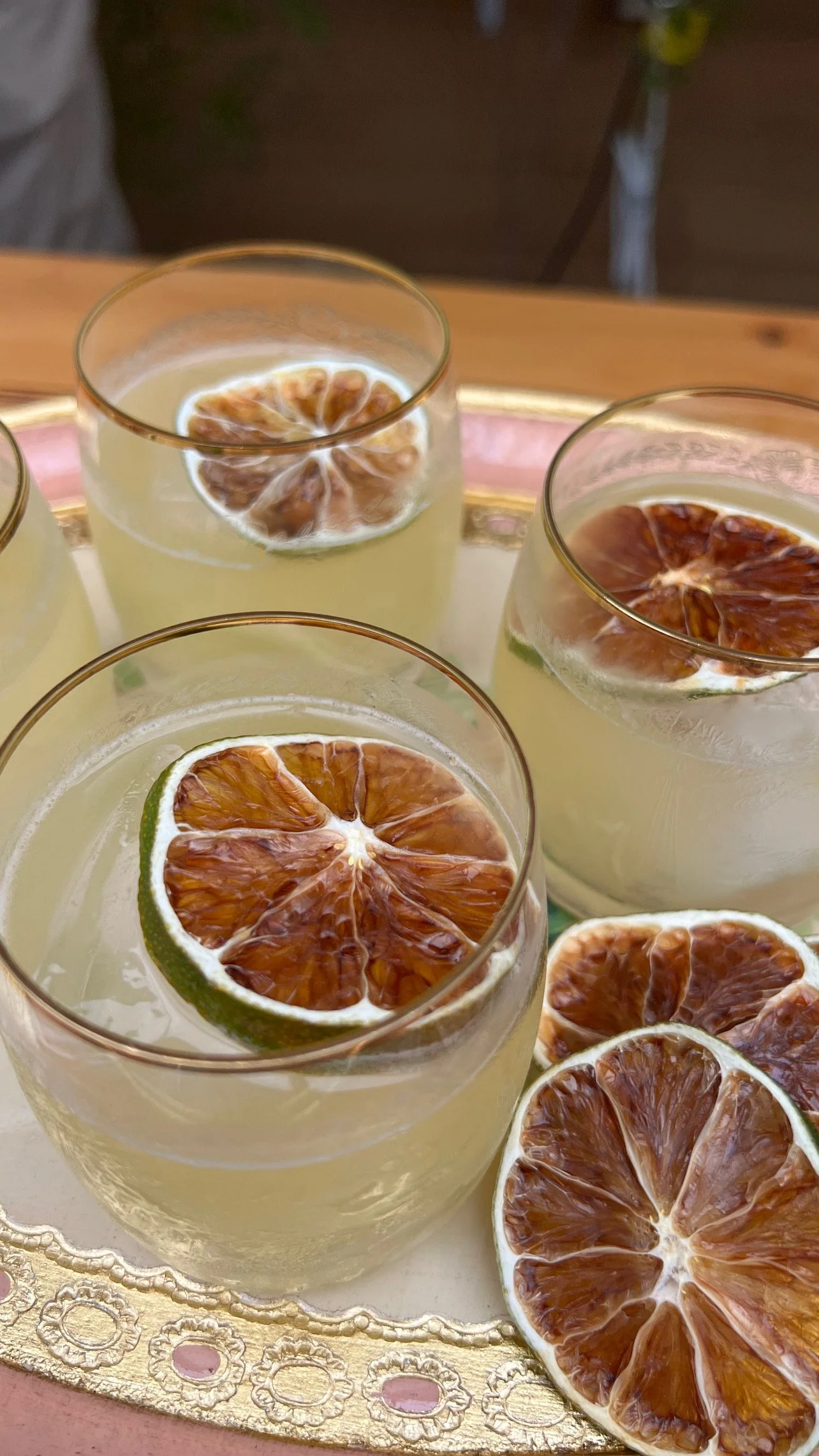 Four glasses of pale yellow cocktail garnished with dried citrus slices, placed on an ornate tray.