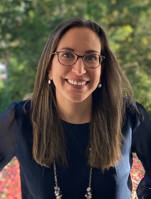 A woman with long brown hair, glasses, and earrings is smiling outdoors, with a blurred green and red foliage background.