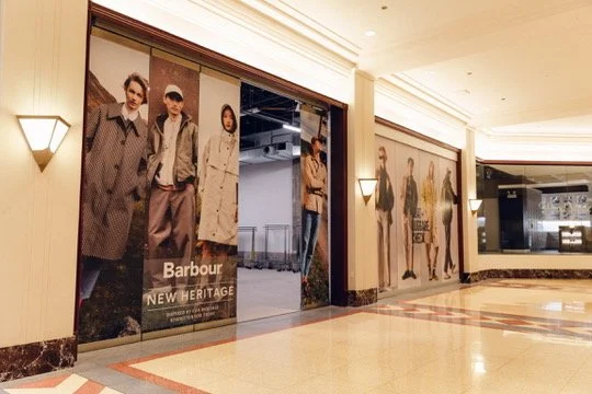 Sign reading 'Coat + Luggage Check' with a woman doing a handstand in a grassy field and mannequins dressed in casual clothing next to a large potted plant in an indoor shopping area.